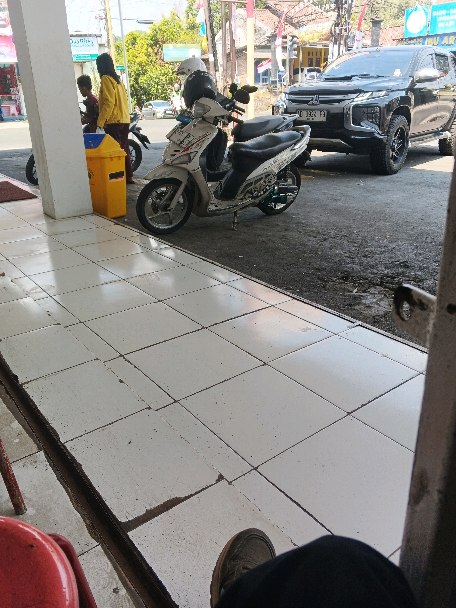 In the city of Genuk Barat, Indonesia, a street scene unfolds with a focus on a parked black SUV. The SUV, identified by the license plate "AD 8924 FB," is positioned on an asphalt surface. In front of the SUV, a white scooter with green accents is parked, its license plate partially obscured. The scooter also has two helmets placed on it, one white and one black.
To the left of the scooter, a yellow trash can is visible. Further left, two individuals are standing near a white pillar. One person, wearing a yellow jacket and dark pants, is looking towards the scooter. The other person, wearing a red shirt, is facing away from the camera. Behind them, a building with signage is partially visible, along with trees and other structures suggestive of an urban or suburban environment. The foreground is dominated by a tiled walkway.
