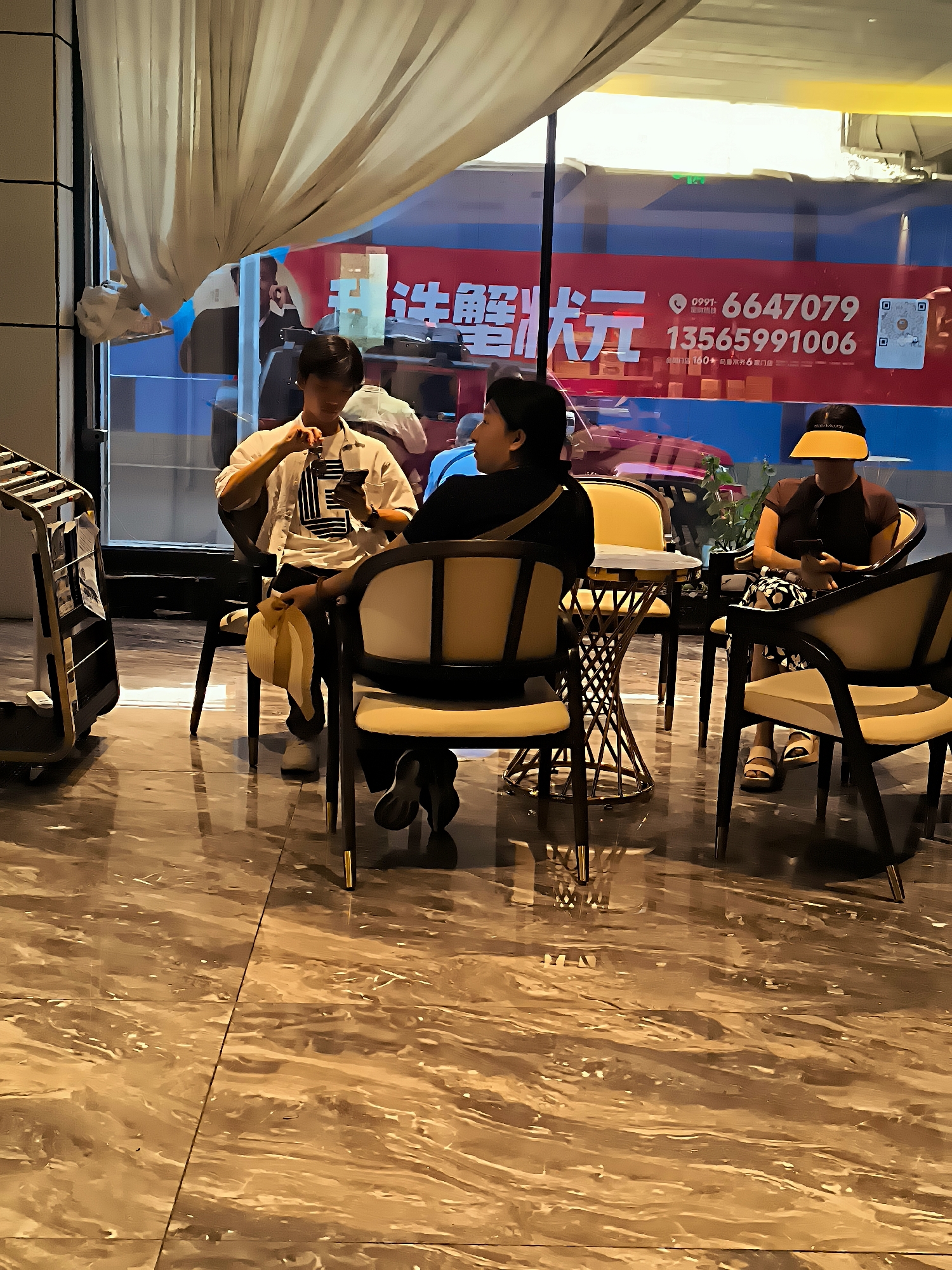 The image depicts a scene inside a cafe or restaurant in Ürümqi, China, with a large window revealing outdoor elements and advertisements. Three individuals are seated at tables. On the left, a young person in a white shirt is looking at their phone, holding a yellow hat in their lap. To the right, a person with dark hair is seated, also looking down at their phone. Further to the right, another person wearing a yellow visor is seated, also engaged with their phone. The interior features modern furniture with chairs upholstered in cream and wood accents, and a distinctive metal-framed table. The floor is polished, reflective marble. Through the window, a bright red advertisement with Chinese characters and phone numbers is visible, along with reflections of the interior and possibly passing vehicles. A luggage cart is partially visible on the far left. The lighting suggests an indoor environment with ambient light and potentially some artificial illumination contributing to the reflections.