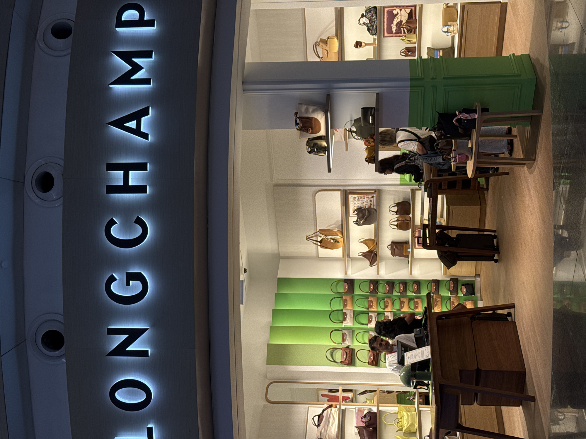 This is a photograph of the exterior of a Longchamp store, likely located in Arnavutköy, Turkey, as indicated by the presence of the brand's prominent illuminated signage. The sign displays the word "LONGCHAMP" in large, backlit capital letters, with a blue-white glow emanating from the edges. Below the sign, the store entrance is visible, revealing an interior display of handbags.
The store's interior is well-lit and features numerous shelves stocked with various styles of Longchamp bags in different colors and sizes. A prominent display wall is covered with a repeating pattern of small, brown and green handbags. Several customers and staff members are visible inside the store. One male employee, wearing a white shirt, is standing behind a wooden counter, appearing to interact with a female customer who is facing away from the camera. Another female customer, also facing away, is examining handbags on a shelf in the background.
The overall impression is one of a high-end retail environment, showcasing the brand's products in an organized and attractive manner. The lighting and display suggest a modern and inviting atmosphere. The reflection on the floor indicates the photograph was taken from a slightly elevated position, possibly from across a walkway or in a mall setting.