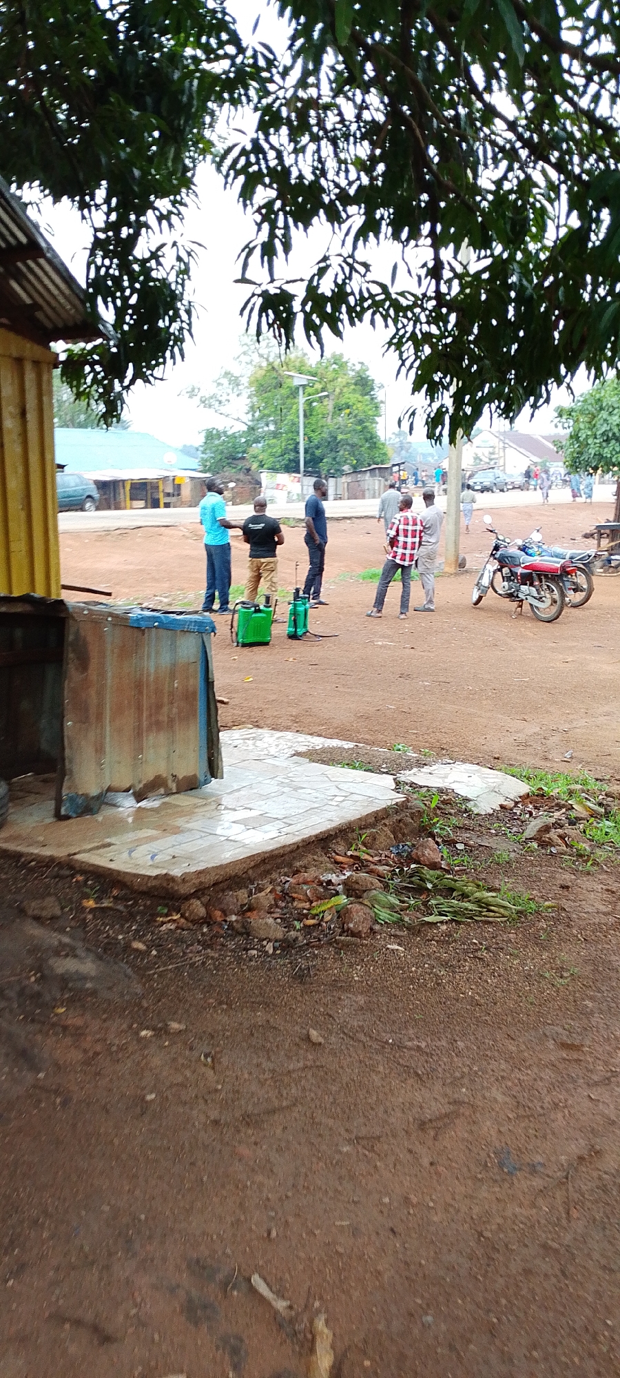 In Gumel, Nigeria, a group of men stand in an outdoor, dusty area near a small building with a yellow and corrugated metal exterior. Two green spray containers are visible on the ground in front of some of the men. Two motorcycles are parked nearby. The sky is overcast, and trees with dark green leaves hang over the scene. In the background, more buildings and vehicles can be seen, suggesting this is a public or commercial space.