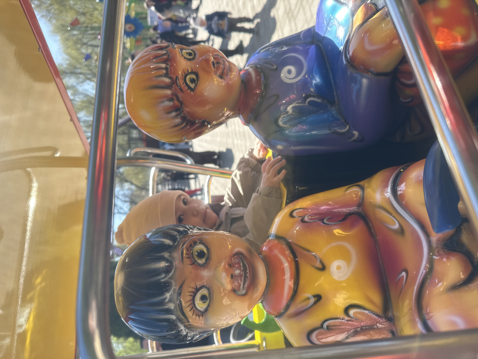 The image shows a child riding a carnival ride in what appears to be a public park in Yekaterinburg, Russia. The child is wearing a beige jacket and beanie and is seated among carousel figures painted to resemble dolls. Other park patrons are visible in the background. The ride appears to be outdoors in daytime, as suggested by natural lighting.