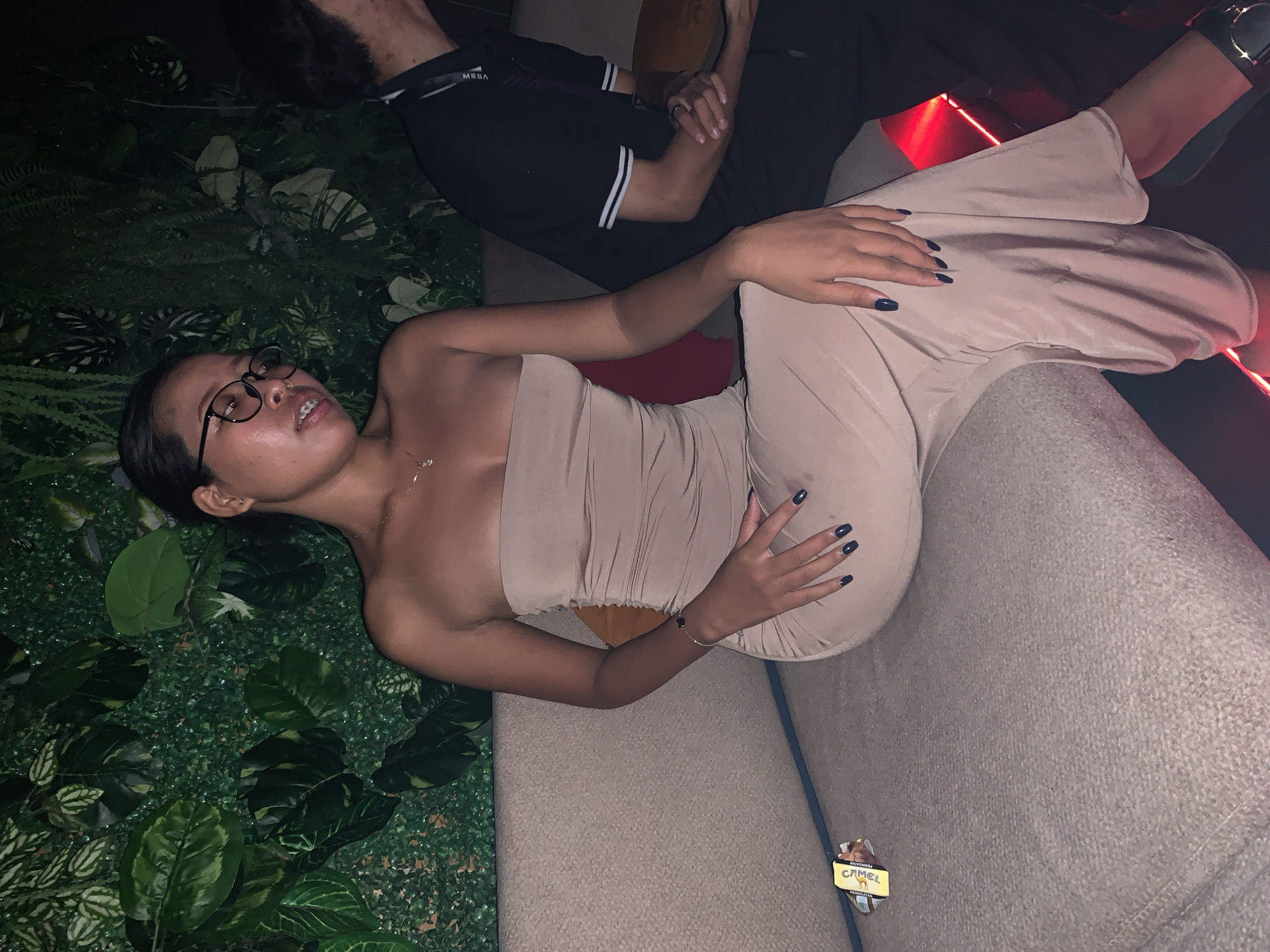 The scene shows a woman reclining on a gray couch, positioned against a wall covered in artificial plants. She wears a strapless tan dress and glasses and has her hands resting gently on her body. A necklace is visible around her neck. Next to her, a person in a black shirt with white stripes sits. A cigarette pack lies on the couch near the woman. The location is North Kuta, Indonesia.
