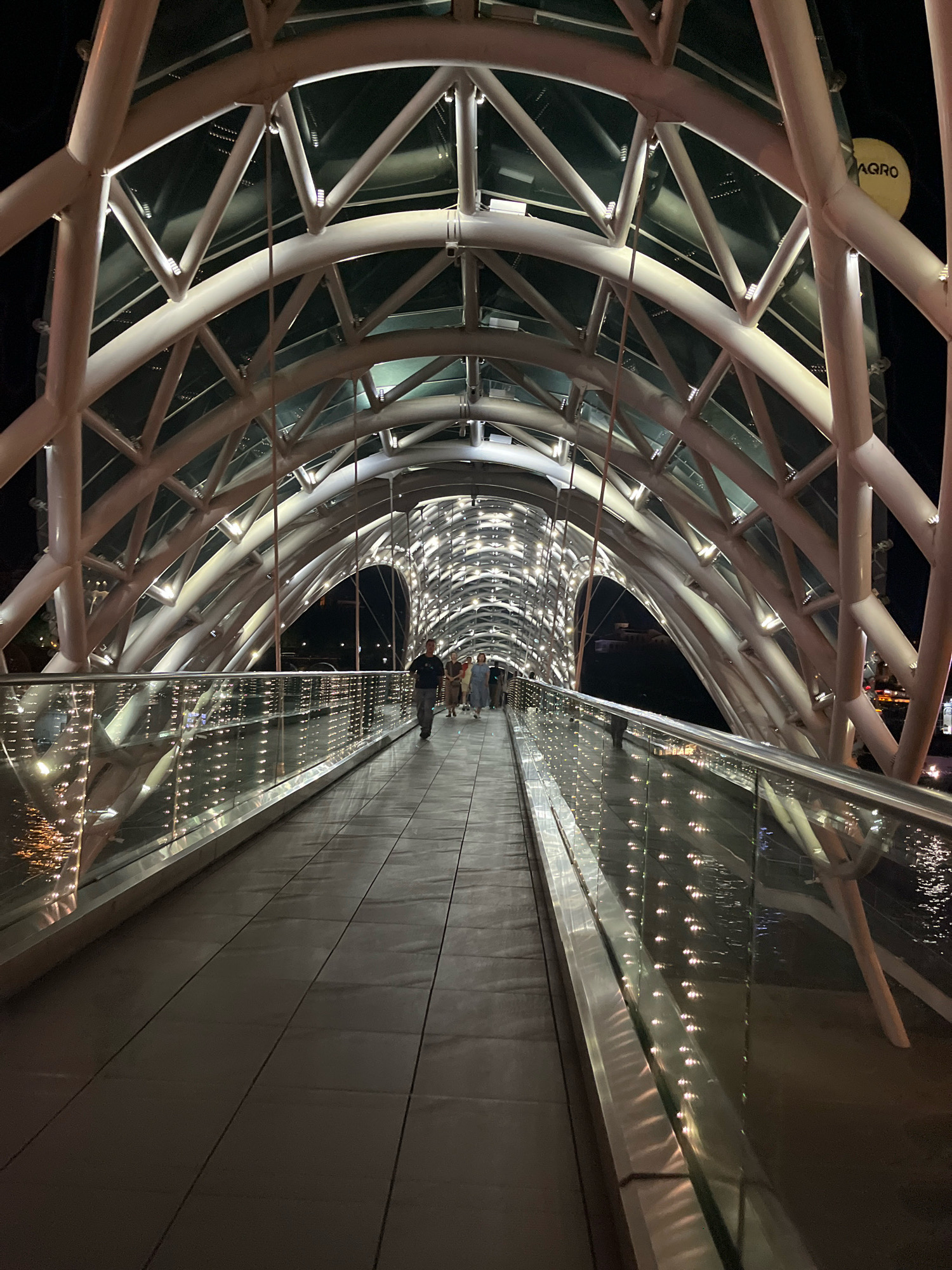 The image captures a nighttime view of the Bridge of Peace in Tbilisi, Georgia. The pedestrian bridge, characterized by its modern, curved architecture with a steel and glass structure, is illuminated by numerous lights that create a vibrant and futuristic ambiance.
The foreground shows the walkway of the bridge, paved with dark tiles and bordered by a sleek glass railing. The glass reflects the lights from the bridge's canopy and the water below, creating a shimmering effect. On the left side of the walkway, the water of the Kura River is visible, reflecting the golden lights.
In the mid-ground, a small group of people are walking across the bridge. They appear to be tourists or locals enjoying a nighttime stroll. Their figures are silhouetted against the bright lights, adding a sense of scale and human presence to the architectural marvel. The people are dressed casually, suggesting a relaxed atmosphere.
The canopy of the bridge, a defining feature, is constructed from a lattice of white steel beams supporting a glass roof. The beams are accented with embedded lights that cast a warm glow, illuminating the pathway and the surrounding area. The design of the canopy creates a tunnel-like effect, drawing the viewer's eye towards the vanishing point in the distance.
In the background, the dark night sky is visible, contrasting with the brightly lit bridge. On the right side of the image, a circular yellow sign with black text is partially visible, reading "AGRO". This sign is likely an advertisement or part of the bridge's surrounding urban landscape. The overall mood is one of awe and wonder, highlighting the bridge as a prominent landmark and an architectural achievement. The time of day is clearly night, and the weather appears to be clear, allowing for a well-lit view of the city.