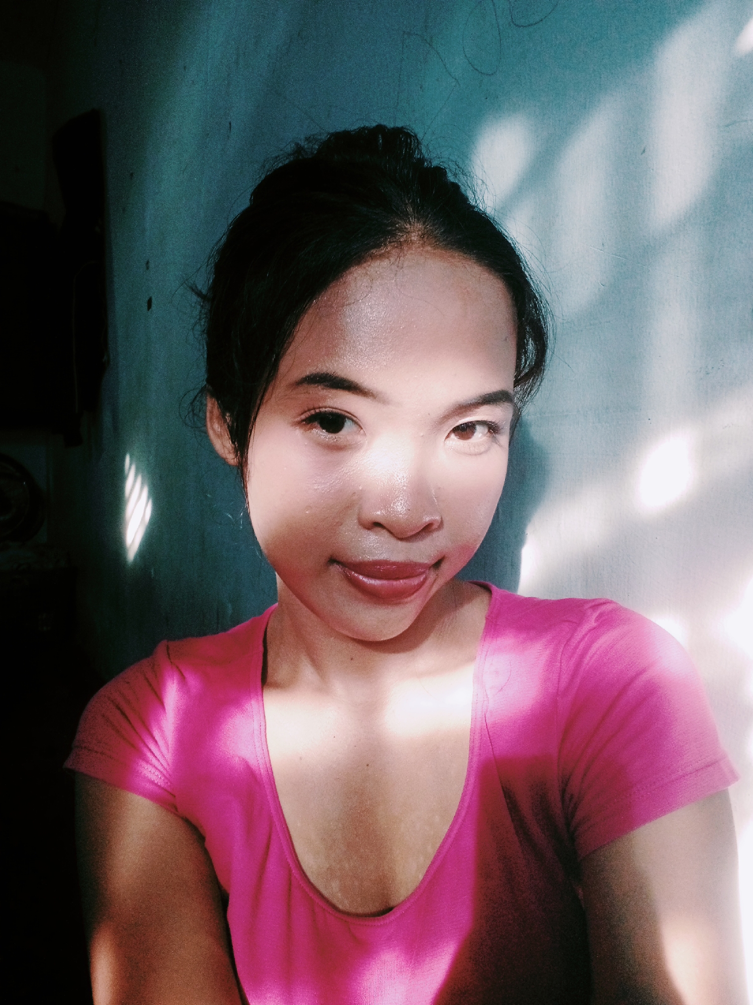A young woman is captured in a close-up selfie, her face angled slightly towards the camera. She has her dark hair pulled up, with a few strands loose around her forehead. Her eyes are open and looking directly at the viewer. She has a subtle smile and her tongue is visible as it lightly touches her lower lip. Her skin appears dewy and flushed, possibly from perspiration or makeup. The lighting in the image is dramatic, with strong sunlight casting shadows and highlights across her face and the wall behind her, suggesting it is daytime and possibly a warm environment. She is wearing a bright pink t-shirt with a scooped neckline. The background is a textured wall, with distinct patterns of light and shadow suggesting it is near a window or opening. The overall mood is intimate and natural. There is no visible text in the image.