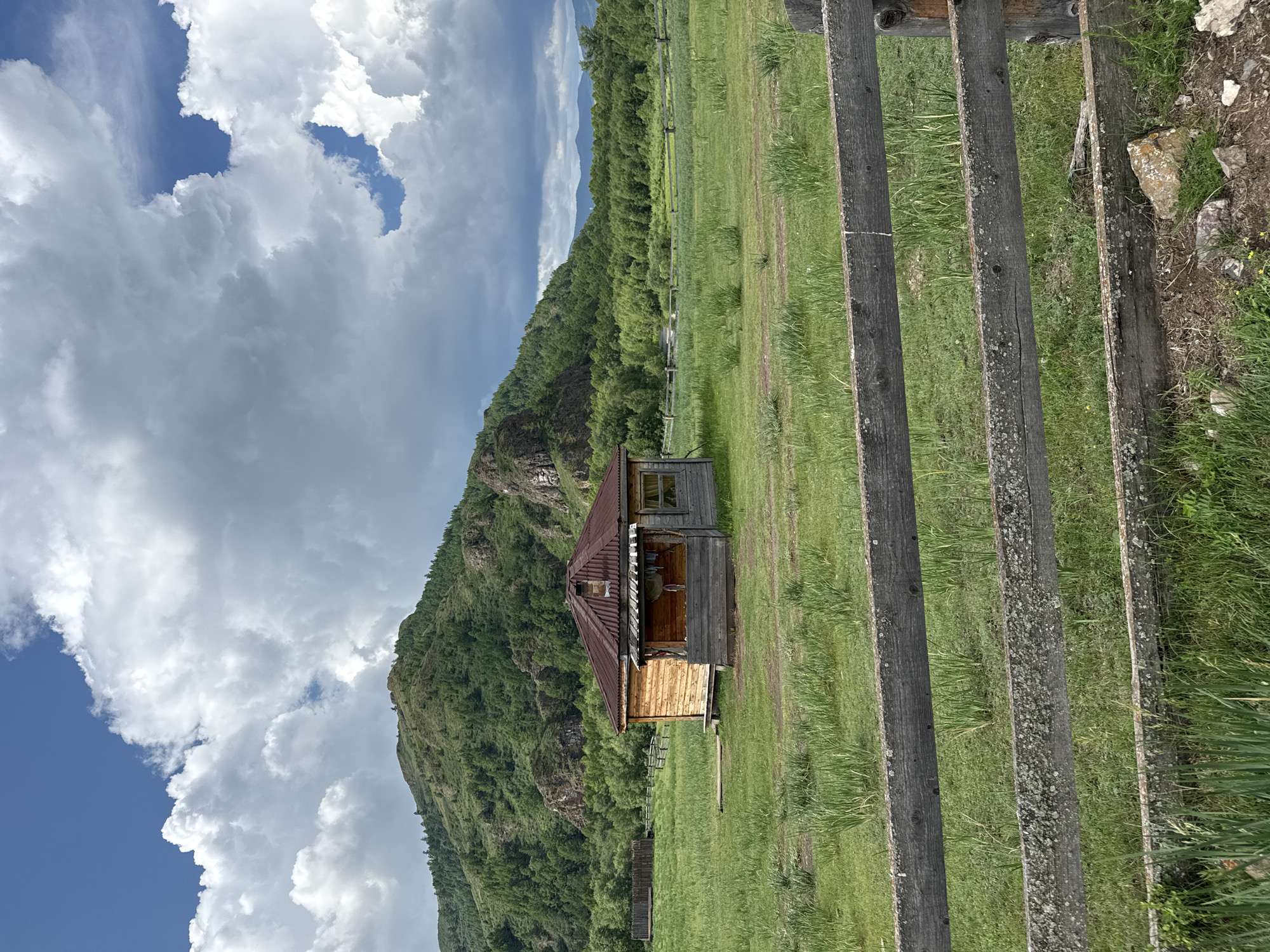 An outdoor, eye-level, medium shot captures a rustic wooden cabin nestled in a grassy field, with a lush, green mountainous backdrop. The sky is a dramatic mix of blue patches and billowing white clouds. In the foreground, a weathered wooden fence, composed of horizontal planks, runs across the bottom of the frame, partially obscuring the view of the field.
The cabin, located in the village of Kazanovka, Russia, is constructed from light-colored wooden logs. It features a prominent, sloping roof made of corrugated metal, with a small chimney. A covered porch extends from the front of the cabin, with a few items of clothing hanging on a line. The cabin's windows are visible, with one appearing to have a glass pane. The surrounding field is covered in tall, green grass and some scattered vegetation.
Behind the cabin, a wooden fence, similar in style to the one in the foreground, encircles a portion of the field. Beyond the fence, the landscape rises into a verdant, tree-covered mountain. The mountain slopes are densely packed with various shades of green foliage, with rocky outcrops visible. The overall scene evokes a sense of rural tranquility and natural beauty.