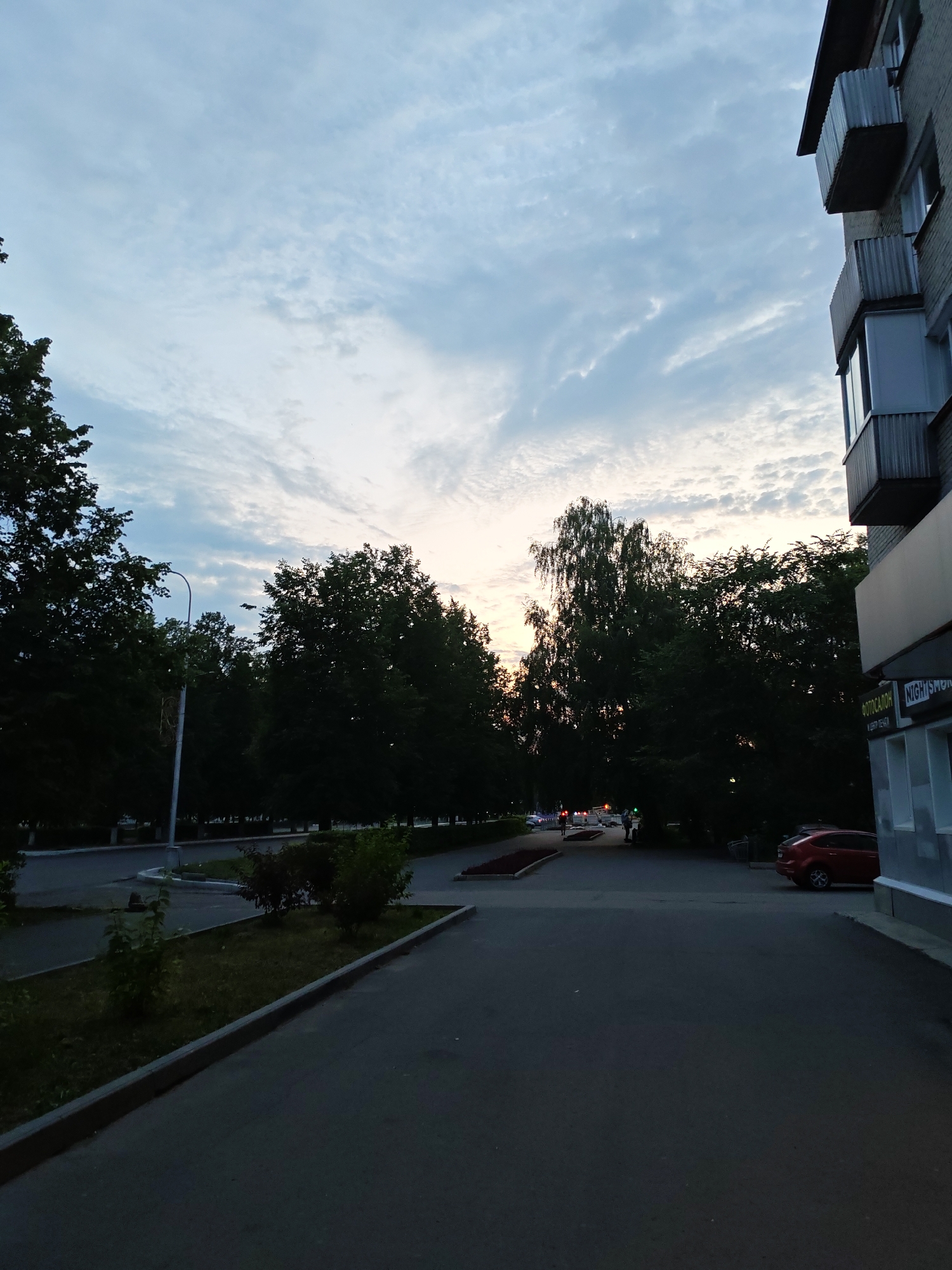 The image shows a street scene in Seversk, Russia, likely during twilight. In the foreground, there is an asphalt road with a concrete curb. To the left, a patch of grass with small bushes is visible. Further left, a lamppost stands beside a row of dark green trees. The background features more trees and a multi-story building on the right side of the frame. The building has balconies with metal railings and a sign that reads "ФОТОСАЛОН" (Photo Salon) and "NIGHT SM" which likely indicates a shop. A red car is parked near the building. In the distance, several cars are visible on the road, and two individuals appear to be walking. The sky is filled with wispy clouds, suggesting the sun is setting or has recently set, casting a soft, diffused light on the scene.