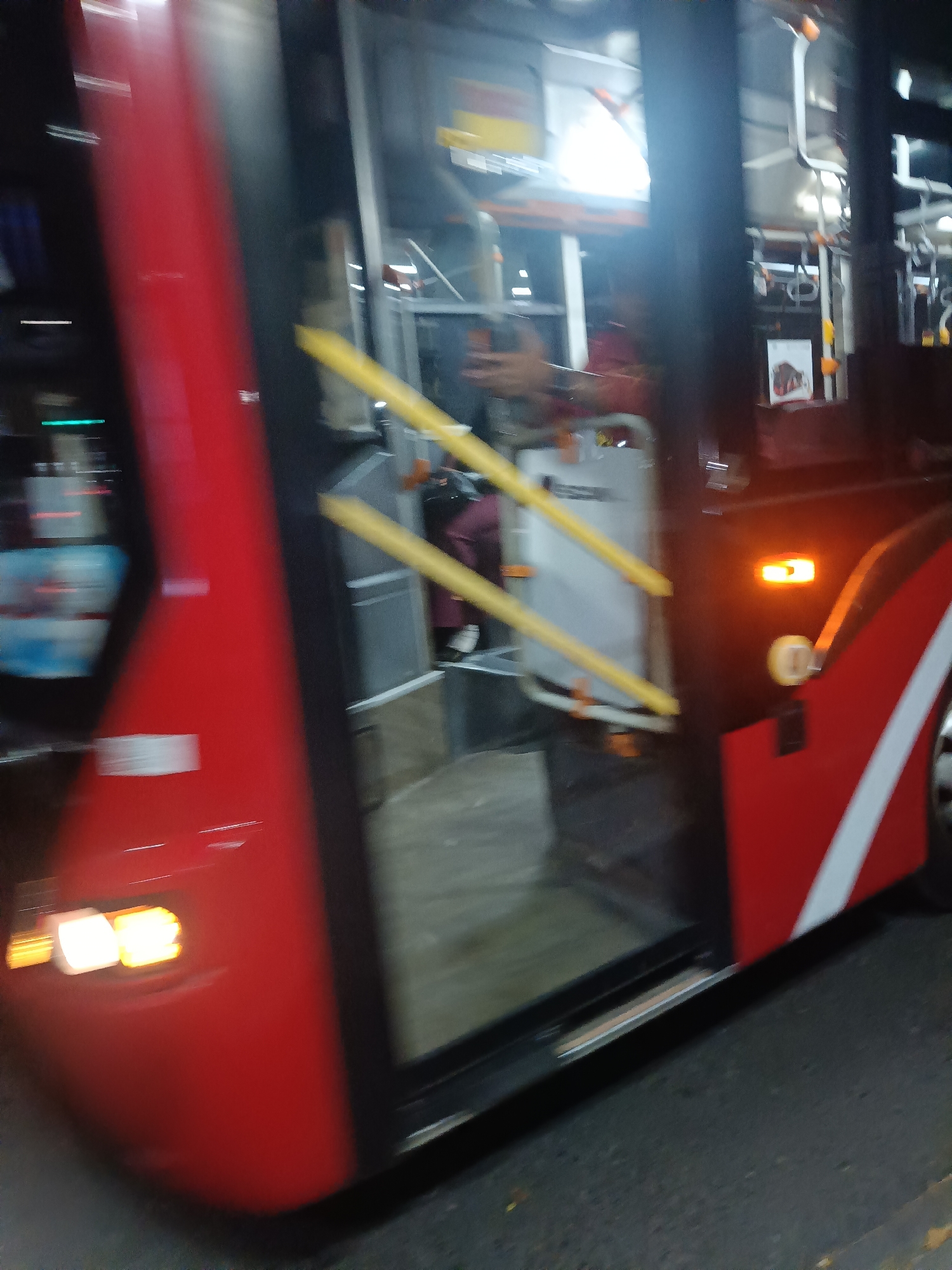 The image shows the side of a red and white bus, likely in Surabaya, Indonesia. The bus has a door that is partially open, revealing a glimpse of the interior. Yellow handrails are visible inside the bus, along with some seats and what appears to be a person in a dark jacket. The exterior of the bus has bright orange indicator lights. The bus is parked on a street, with asphalt visible on the ground. The overall scene is slightly blurred, suggesting movement or a low-light environment.