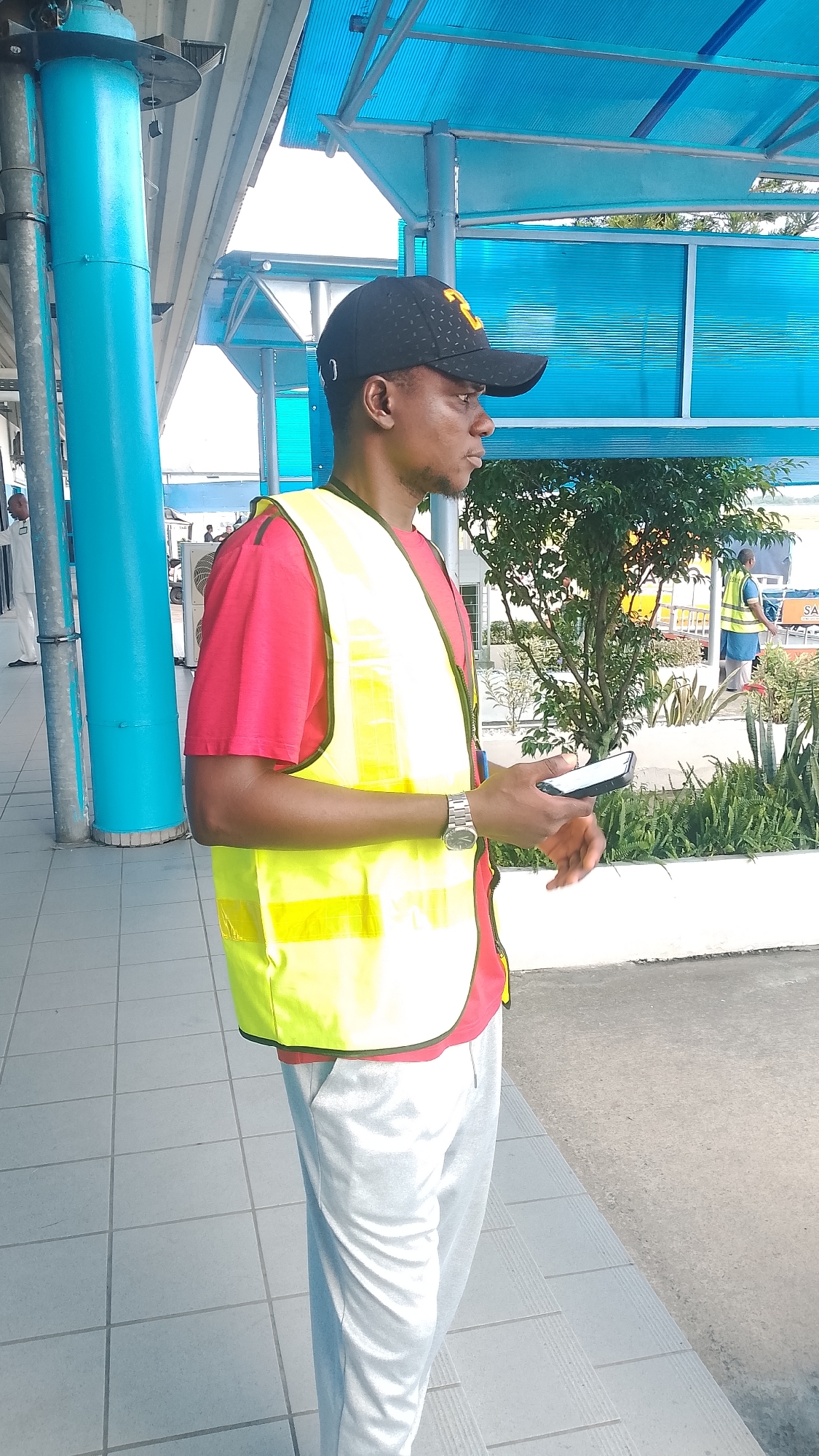 A man, wearing a red t-shirt, a yellow high-visibility vest, and a black baseball cap, stands on a tiled walkway. He is holding a smartphone in his right hand and looking to his right. Behind him, a blue awning covers the walkway, supported by a teal-colored pillar on the left. In the background, there are trees and other people, one of whom is also wearing a yellow high-visibility vest. The scene appears to be an outdoor public area, possibly a bus station or a similar transportation hub, in Orerokpe, Nigeria. The lighting suggests it is daytime, with bright sunlight.