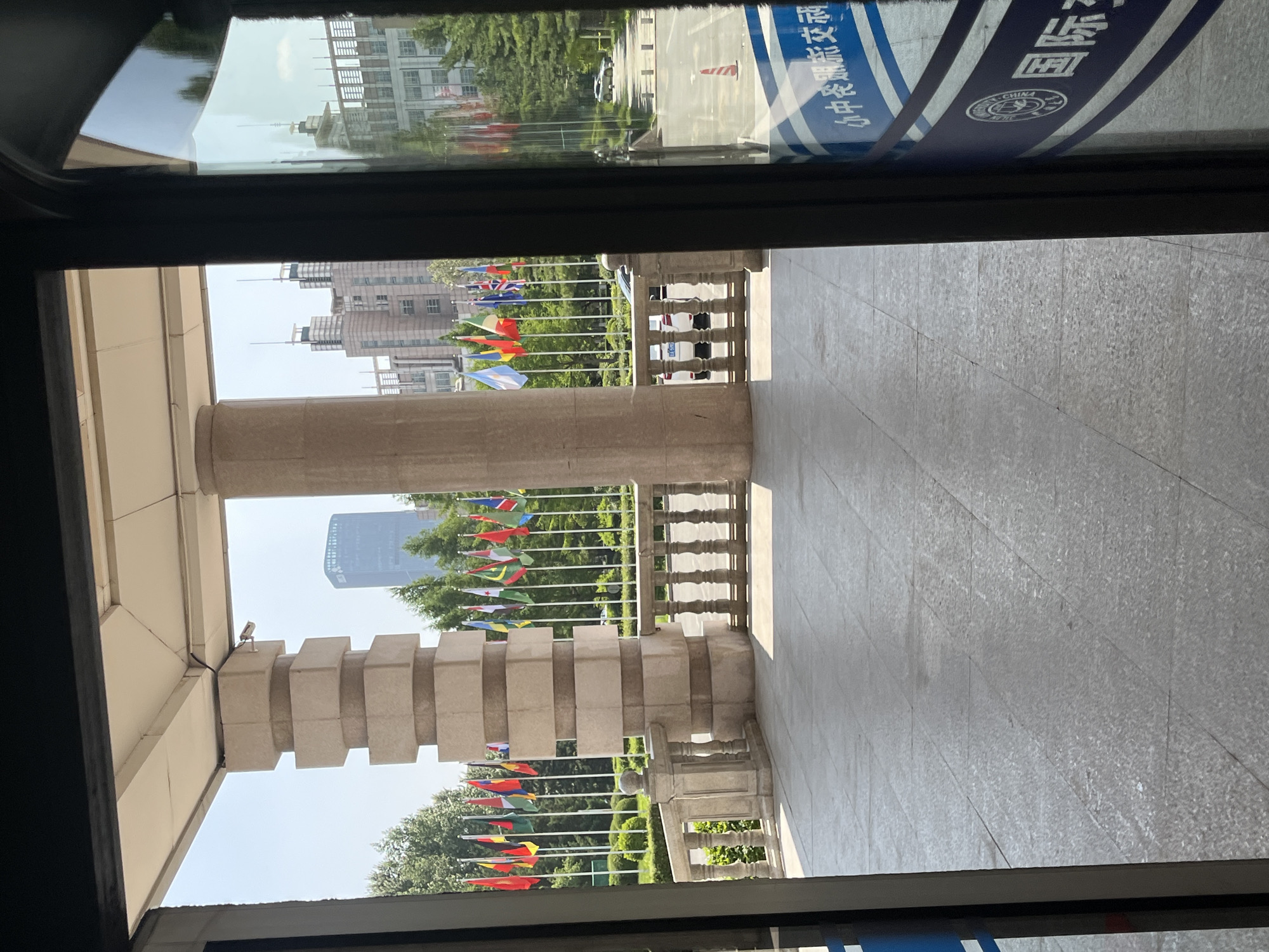 The image depicts an outdoor scene viewed from within a building, likely an entrance. In the foreground, a paved walkway is visible, with a large, segmented stone pillar on the left and a stone balustrade stretching to the right. Beyond the balustrade, a row of national flags on poles is displayed, set against a backdrop of trees and modern buildings. One prominent skyscraper with a distinct architectural design is visible in the distance, as are several other multi-story buildings. Two white cars are parked on a road behind the flags. The overall environment suggests a formal or governmental setting, potentially in a city park or a plaza. On the right side of the image, a glass door or window reveals reflections of the surroundings and a blue and white sign with Chinese characters and a circular emblem, suggesting this is a location in China. Based on the presence of the signage and architectural style, the scene is likely located in Changchun City, China, given the visual cues of urban development and potential for international representation with the flags. The image is captured during daylight hours under a clear sky. No people are discernible in the scene.