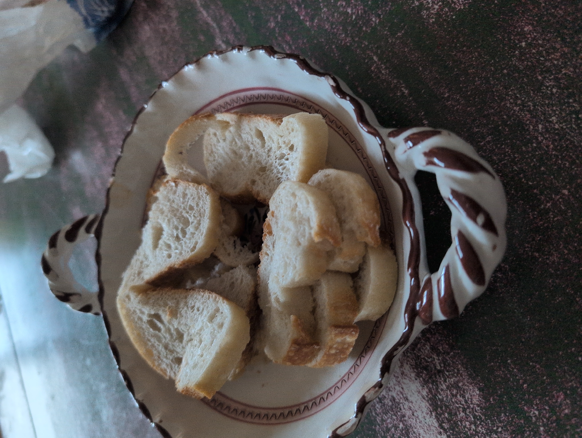 A close-up, overhead shot shows a white ceramic dish with brown twisting handles filled with sliced bread. The dish has a rim decorated with a red zigzag pattern. The bread slices are round and appear to be from a baguette. The dish rests on a dark, speckled surface, possibly a table. In the upper right corner, a blurred white object, possibly a napkin or paper, is visible. The lighting suggests an indoor setting, and the focus is on the food and dish. There are no people or animals present in the image. The location in Harsewinkel, Germany, is not discernible from the visual information provided.