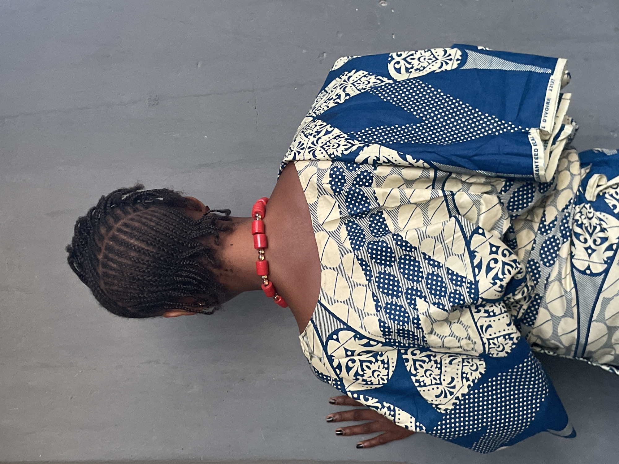 A woman with braided hair kneels on a gray surface wearing a blue and white patterned garment and a red beaded necklace. Her hands are placed flat on the ground in front of her. The printed text "E D'IVOIRE 22127" is visible on the fabric. The location is unspecified, but the presence of the fabric roll indicates Badagry, Nigeria.