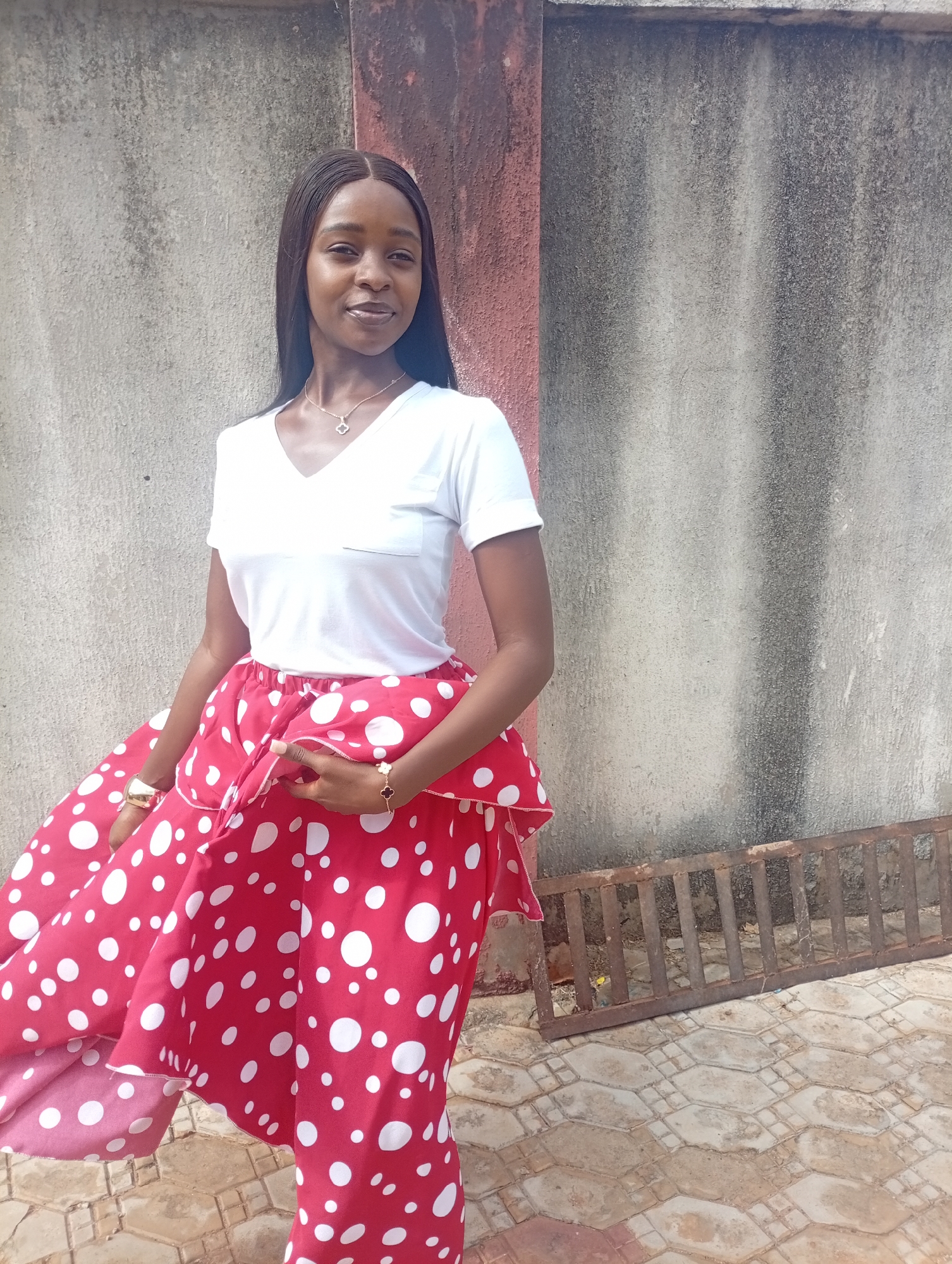 A woman stands in front of a textured, grey concrete wall with a rust-colored vertical section to the left. She wears a white v-neck t-shirt and a red skirt with large white polka dots. The skirt's fabric is partially lifted by her hand, revealing a pink underside. She wears a gold watch on her left wrist and a clover-shaped charm bracelet on her right. The woman has long, dark hair and is smiling. A metal grate lies on a tiled surface near the bottom right of the image. The scene is in Rido, Nigeria.