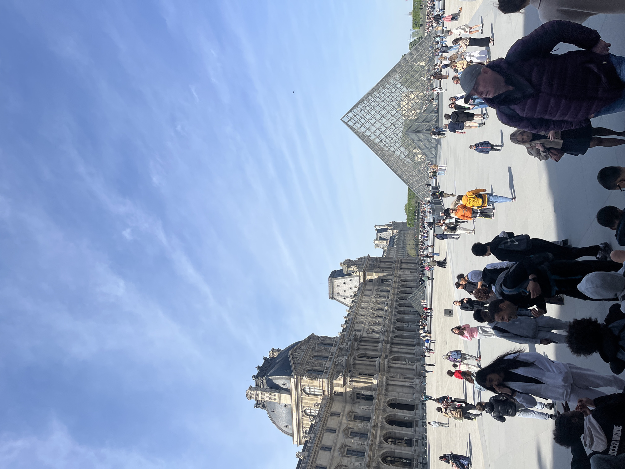 The image shows the Louvre Museum and its iconic glass pyramid in Paris, France. A large crowd of people is milling around the plaza. The architecture of the museum is visible in the background. The sky is blue with light clouds.