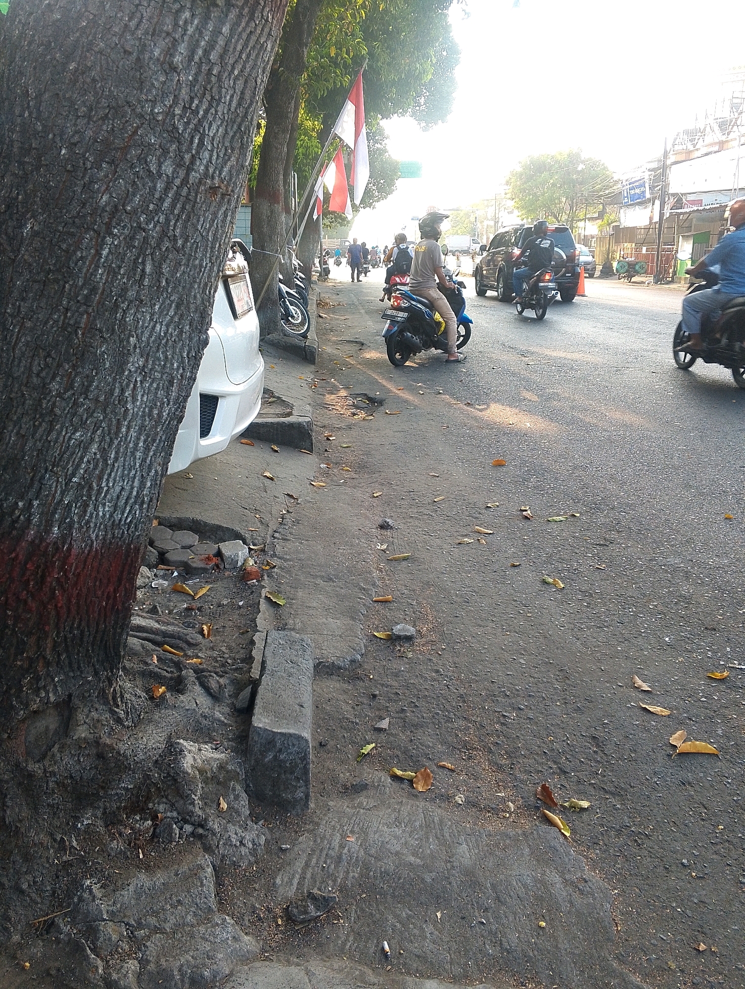 The image depicts a street scene with heavy traffic and pedestrians. A large tree trunk dominates the left foreground, with visible red markings on its base. The sidewalk, in front of parked motorcycles and a white car, is cracked and uneven, with debris and fallen leaves scattered on it.
In the background, several motorcycles are in motion, some carrying passengers. A few cars are also present on the road. People are visible riding on the motorcycles and walking on the sidewalk. Two Indonesian flags are attached to poles along the side of the road. The overall environment appears to be a busy urban street bathed in bright, possibly late afternoon sunlight, indicated by the long shadows and the glare from the sky. Buildings and utility poles line the far side of the street.
