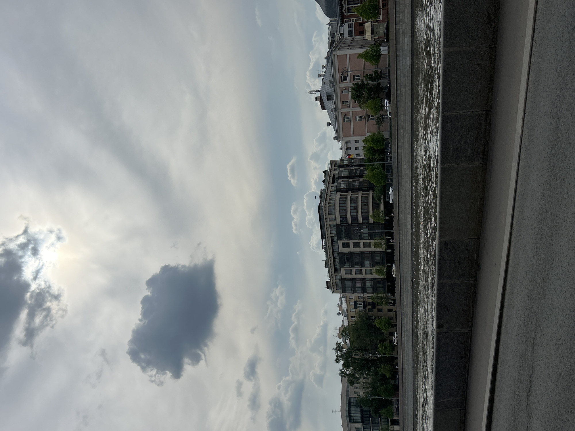 The image shows a cityscape under a cloudy sky. Buildings are visible along the horizon, varying in height and architectural style, some with trees interspersed. Adjacent to the buildings is a body of water, likely a river or canal, with a stone embankment visible along the edge. The right side of the image features a paved walkway or road, indicating an urban environment. The lighting suggests daytime, with the sun obscured by clouds.