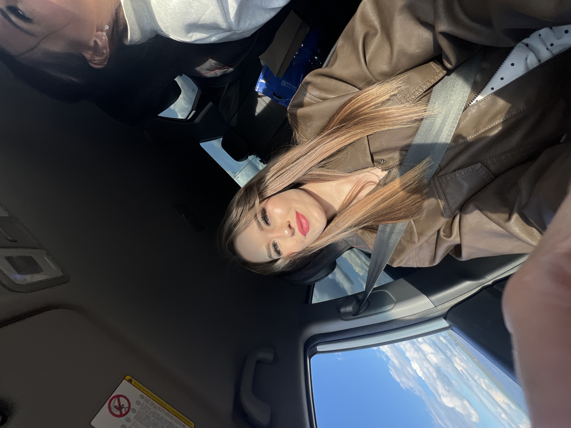 A woman is inside a vehicle, wearing a brown leather jacket and a seatbelt. The sky and clouds are visible through the car window. The location, city of Chaikovsky, Russia cannot be determined from the visual data provided.