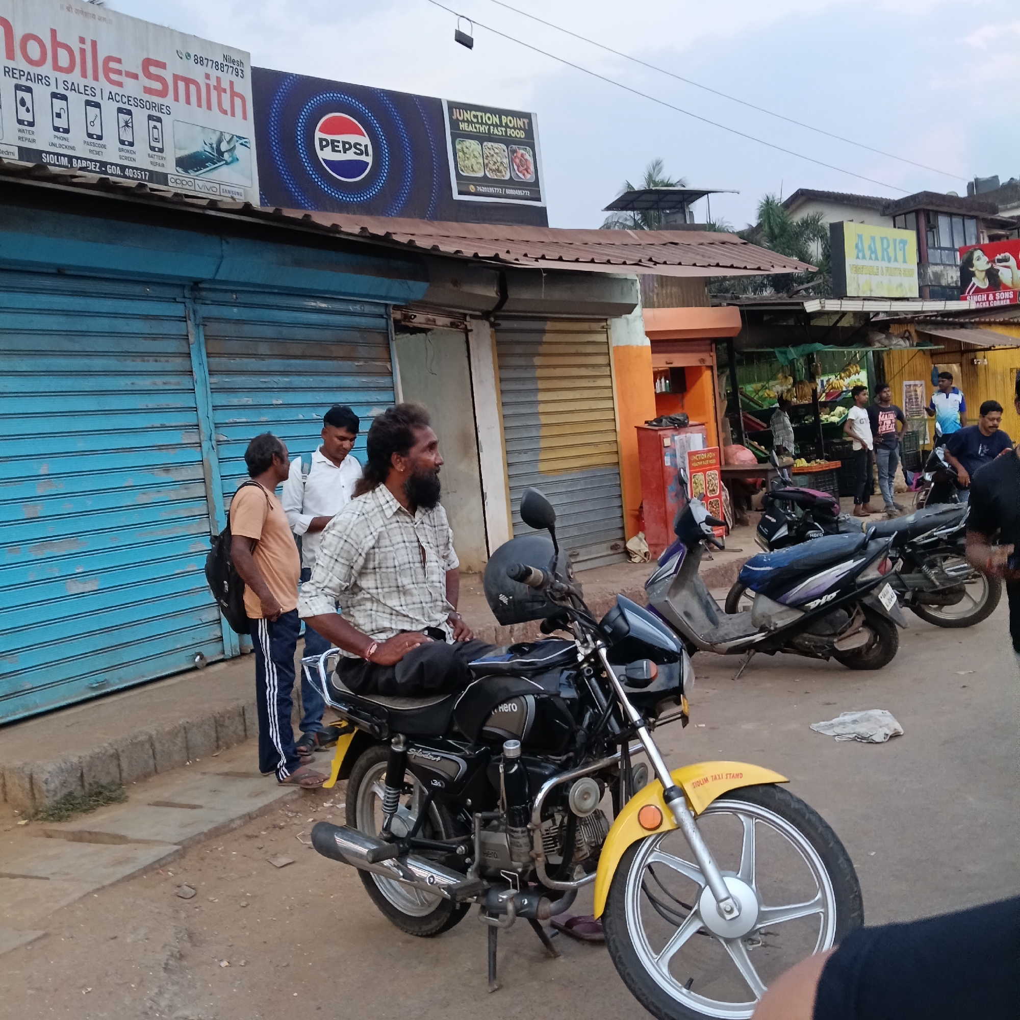 The image depicts a street scene in what appears to be Oxel, India. The primary focal point is a man sitting on a black Hero Splendor motorcycle with a distinctive yellow front mudguard marked "SIOLIM TAXI STAND". He has a full beard and long hair, and is wearing a plaid shirt. Two other men stand near him, one wearing an orange shirt and the other a white shirt.
Behind them is a building with closed blue corrugated metal shutters. Above the shutters is a sign for "Mobile-Smith" offering mobile phone repairs and accessories, next to a "Pepsi" advertisement and a sign for "JUNCTION POINT HEALTHY FAST FOOD".
Further down the street are other shops and people. In the background, there is a partially visible sign for "AARIT" and another sign for "SINGH & SONS". There are also other motorcycles parked along the side of the street. The weather appears to be overcast.