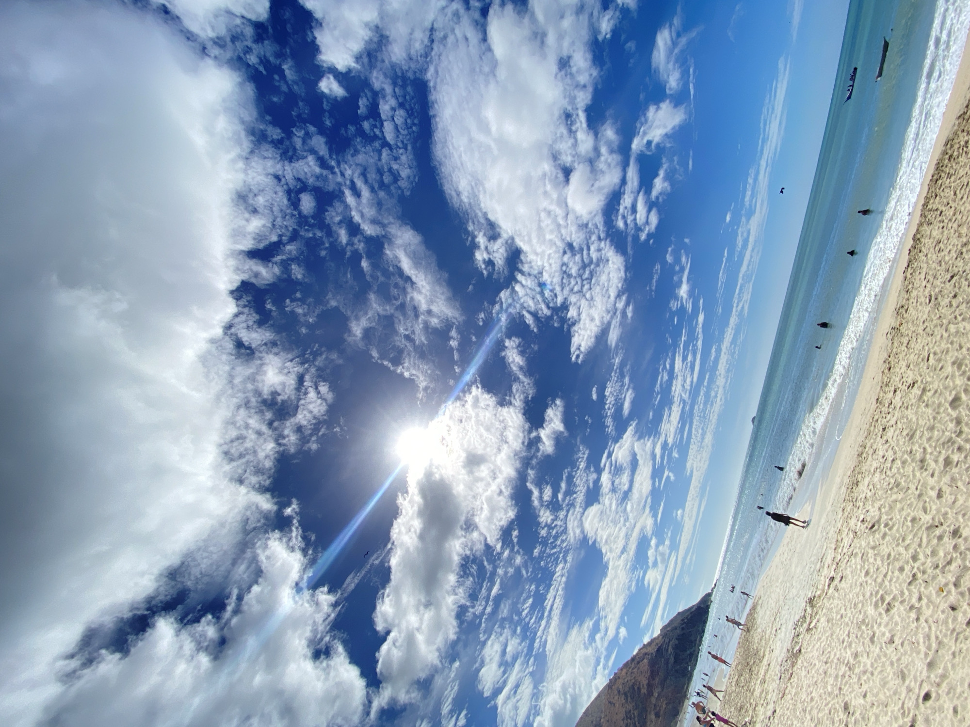 The image shows a beach scene in Pedro González, Venezuela, angled upwards towards a partly cloudy sky. The bright sun is partially obscured by clouds, creating a glare. The beach is sandy with people walking and standing near the shoreline. The ocean is light blue with small waves. In the distance, a boat is visible on the water. A dark land formation is on the left side of the beach. The sky is predominantly blue with scattered white clouds.
