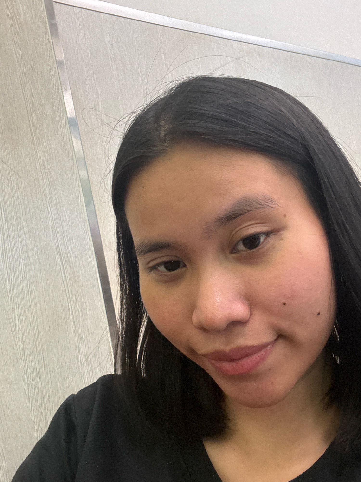 A young woman with long, dark hair is taking a selfie in what appears to be an indoor setting. She is wearing a black top. The background consists of a light-colored wall with vertical wooden paneling and a metallic vertical strip. The image is cropped tightly on her face and upper torso. The location is identified as East Jakarta, Indonesia.