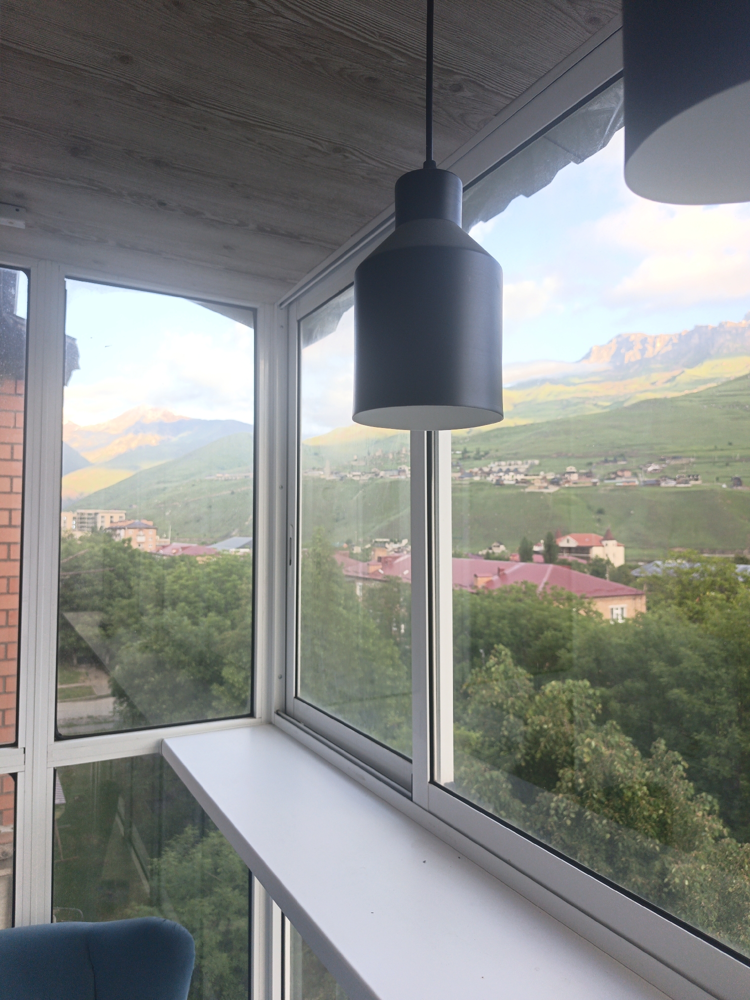This image provides an interior view from a room in Verkhny Fiagdon, Russia, looking out through a large window. The foreground shows a portion of a light-colored windowsill and a dark gray pendant lamp hanging from a wooden-paneled ceiling. The window reveals a panoramic vista of the town and surrounding mountainous landscape.
The town is characterized by scattered buildings with red and orange roofs, nestled amongst lush green trees. In the distance, rolling hills covered in green vegetation rise up to meet imposing mountains, which are bathed in the warm light of what appears to be either sunrise or sunset, casting golden hues on their peaks. The sky above is partly cloudy, with soft white clouds drifting across a pale blue expanse.
The architectural style of the buildings suggests a rural or semi-rural setting typical of a mountainous region. The overall impression is one of tranquility and natural beauty, with the modern interior contrasting with the timeless landscape outside. There are no people visible in the image.