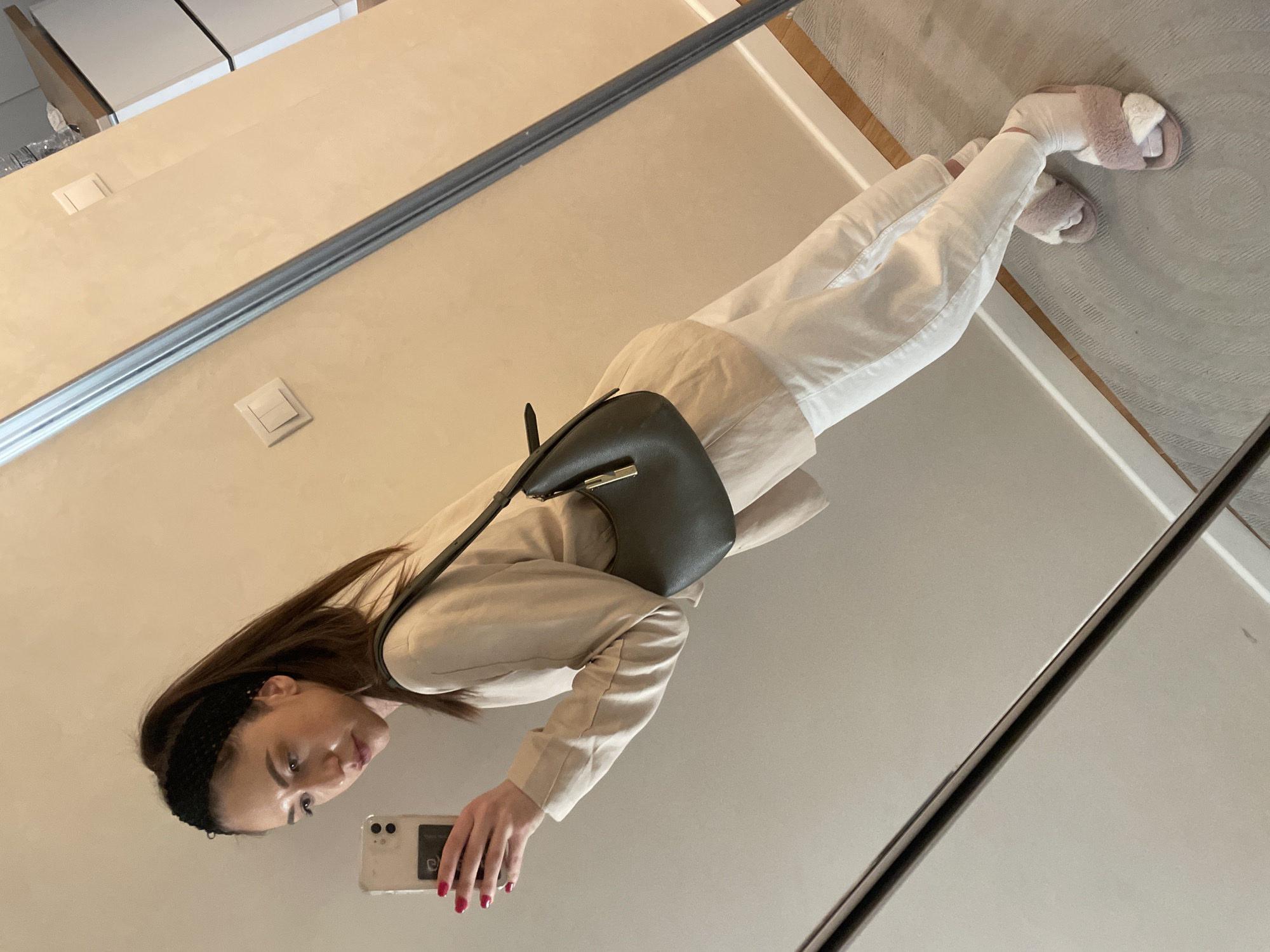 A woman stands in front of a mirror, capturing a self-portrait with her smartphone. She is wearing a light-colored blazer, white pants, and fuzzy slippers. A black purse hangs over her shoulder. The location appears to be indoors, with elements suggesting a residential or dressing room setting. The exact city of Varna, Bulgaria, cannot be determined from the image.