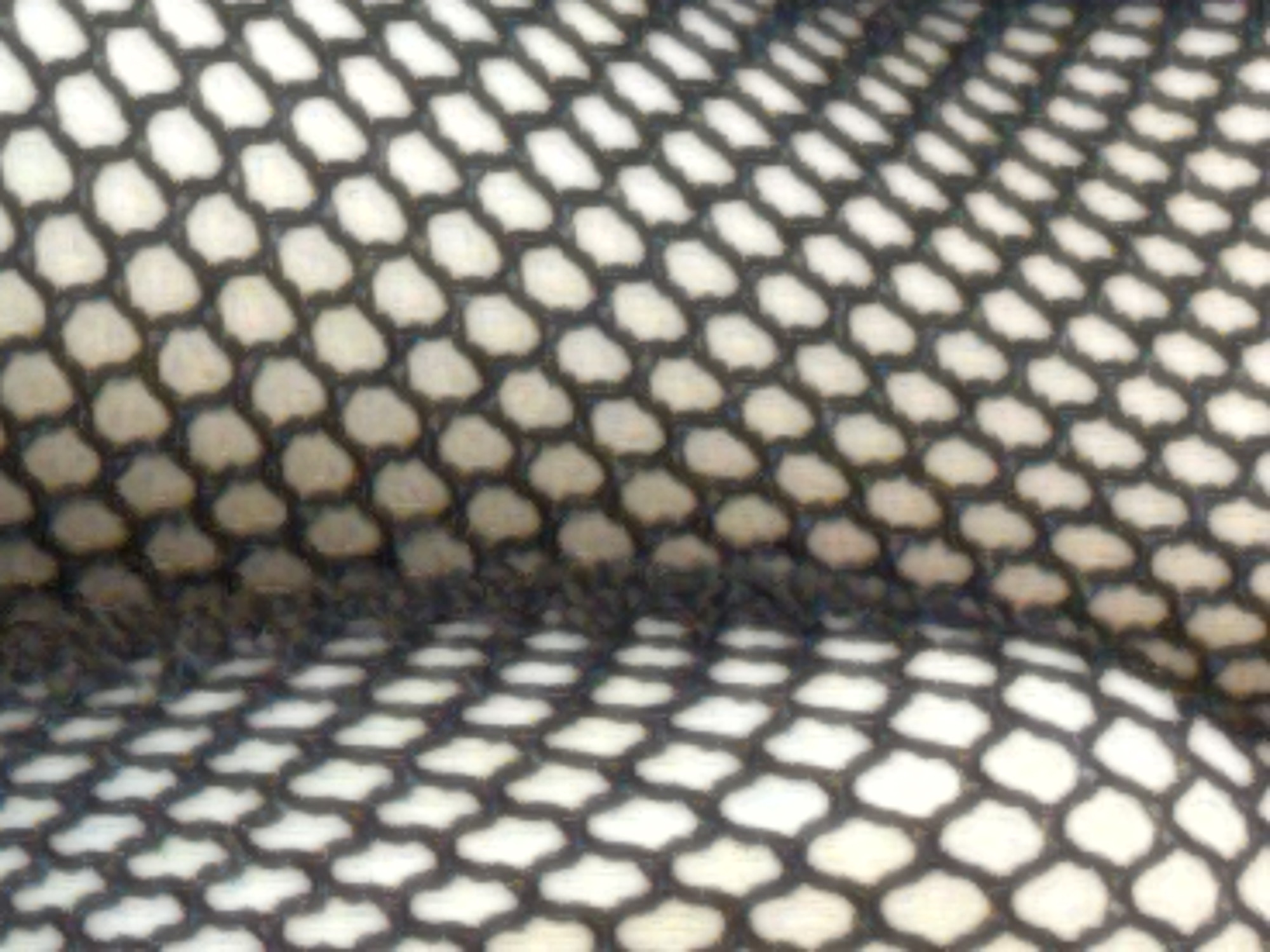 The image shows a close-up, abstract view of a black fishnet material, with a lighter colored material visible through the diamond-shaped openings. The lighting creates a gradient, with the left side appearing brighter and more in focus, gradually becoming darker and more blurred towards the right. The texture of the fishnet is the dominant feature, creating a repeating hexagonal pattern. The image does not contain any discernible elements related to the city of Kholmsk, Russia, or any specific location within it. There are no people, subjects, objects, actions, interactions, or environmental details that can be identified.