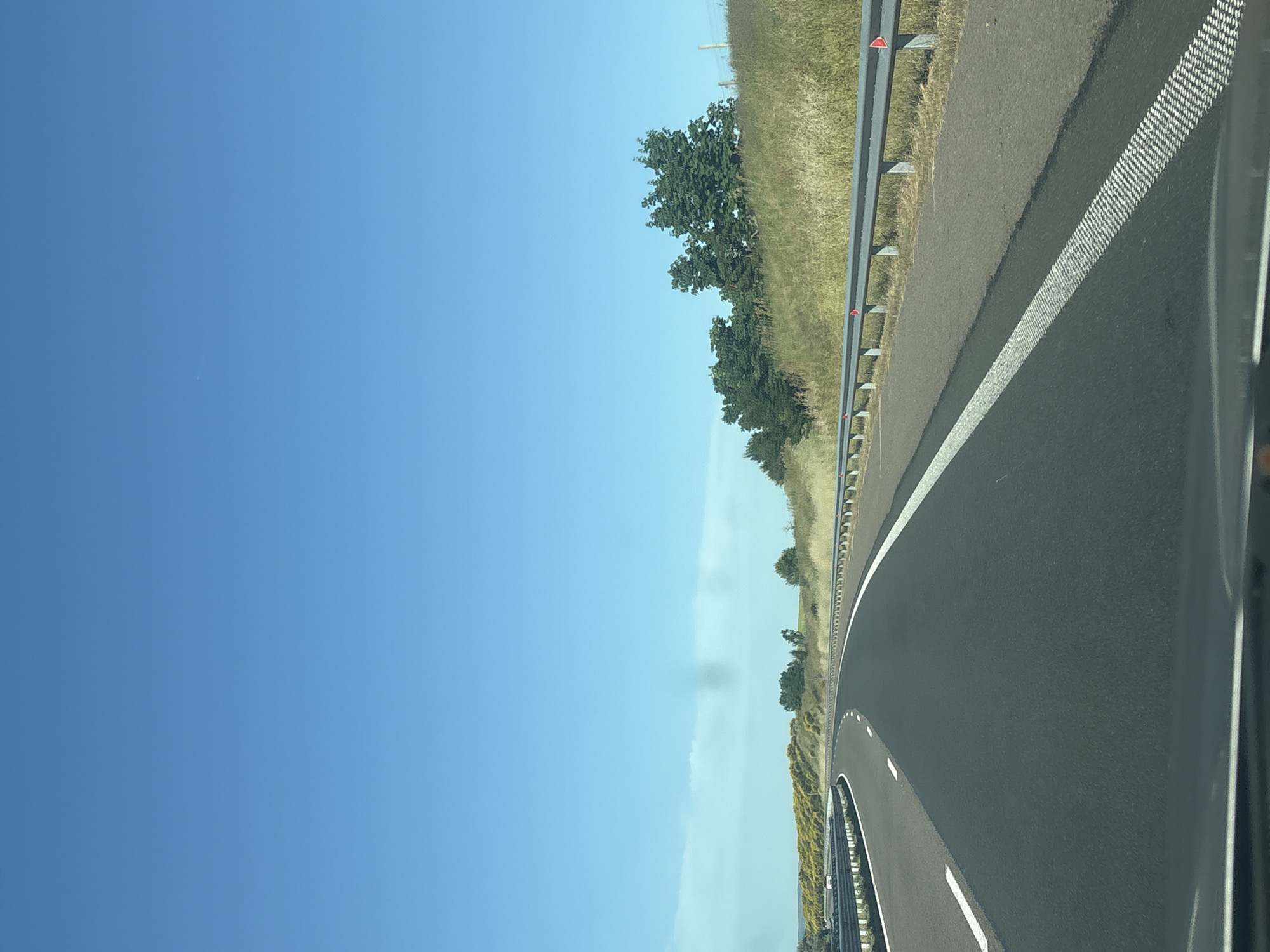 The image shows a clear blue sky over a highway in Vratitsa, Bulgaria. The highway curves to the left, with a metal guardrail on the right side and a dotted white line marking the lane. The surrounding landscape consists of grassy hills dotted with trees and yellow flowering bushes. The road surface appears to be asphalt. There are no people or vehicles visible in the immediate foreground.