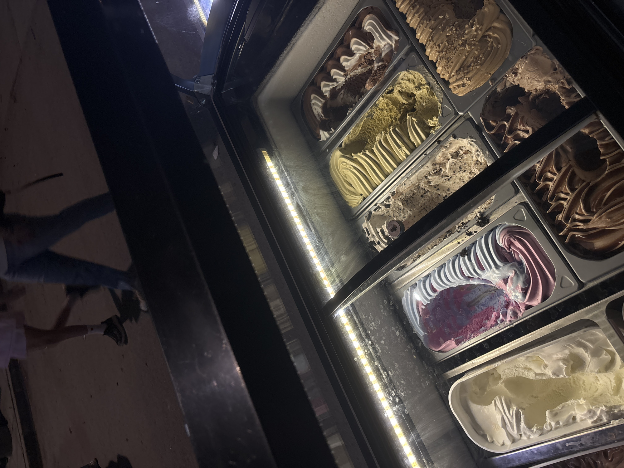 The scene depicts a nighttime view of an illuminated gelato display case in the city of Kostievo, Bulgaria. In the foreground, a black-framed refrigerated display case, likely situated outdoors or in an open-air commercial setting, features multiple trays of artisan gelato. An LED strip light along the top interior of the case brightly illuminates the various flavors. The glass front exhibits some condensation or light smudges.
From left to right, the visible gelato flavors include: a rich, dark chocolate or coffee, a light cream-colored or vanilla, a vibrant pink-purple base with light blue-white swirls, a speckled off-white with dark inclusions resembling cookies and cream, a light green or pistachio, a caramel or praline with visible toppings, and a prominent tray of chocolate and white cream swirled gelato. The gelato is presented in uniform, rectangular metal trays, each with distinct, decorative swirls and peaks from scooping.
In the blurred background, two partial figures of people are visible on a dark, paved surface, suggesting a street or walkway. One figure, on the left, wears light-colored attire and dark athletic shoes with white socks, moving right to left. Another figure, on the right, wears blue jeans and moves left to right. The ambient lighting in the background is dim, contrasting with the bright illumination of the display case.
