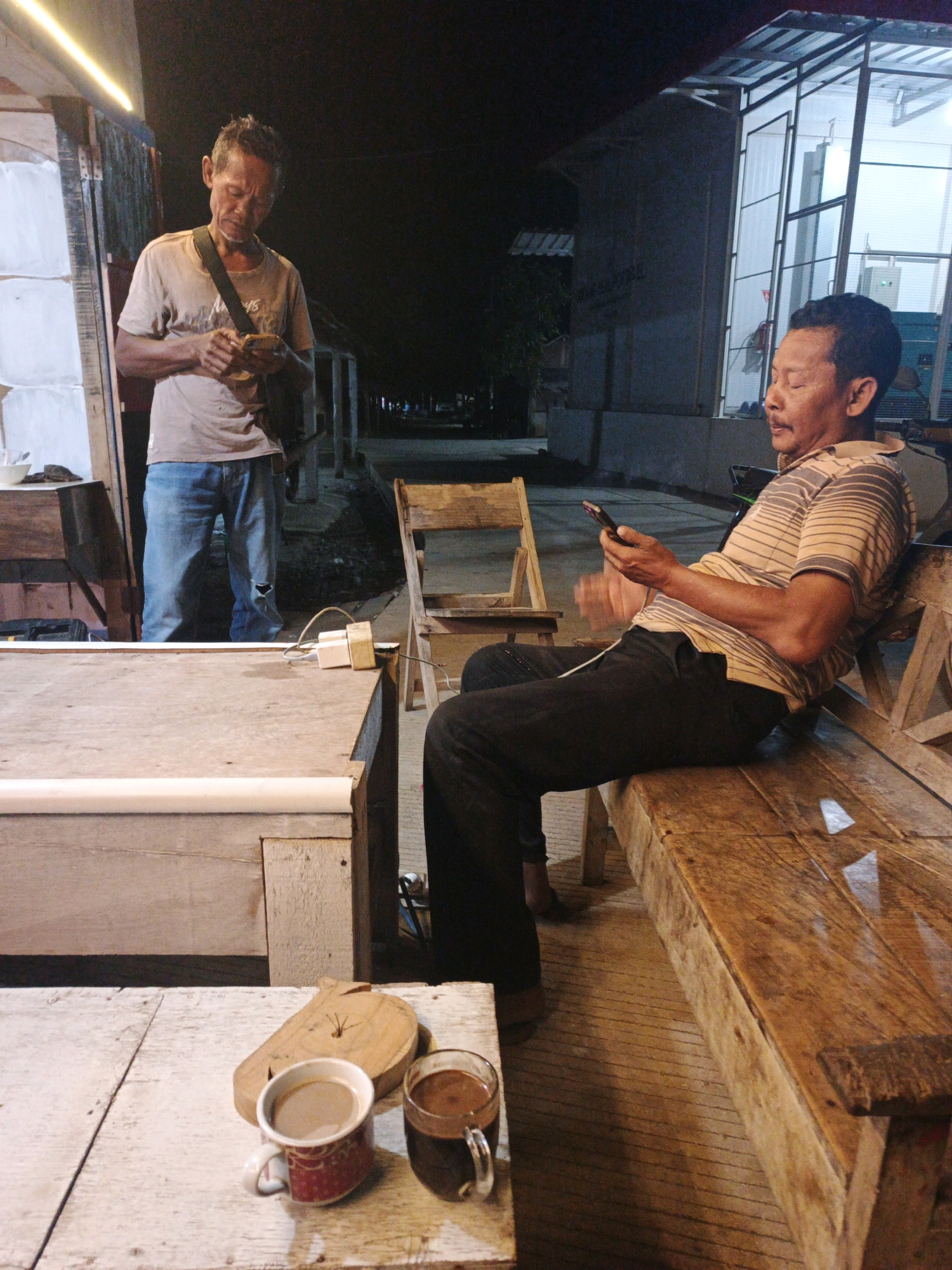 Two men are present in the scene. The man on the left, wearing a light-colored t-shirt and blue jeans, stands and looks down at his phone. He has a bag slung across his chest. The man on the right, in a striped t-shirt and dark pants, sits on a wooden bench, also looking at his phone. A wooden chair is visible behind him. In the foreground, on a wooden surface, sit two cups, one containing a light brown liquid and the other a dark brown liquid, next to a carved wooden object. The scene appears to be an outdoor setting at night, possibly a roadside stall or eatery, with a building and some lighting visible in the background. The environment suggests a casual, perhaps informal, gathering place in Indonesia.