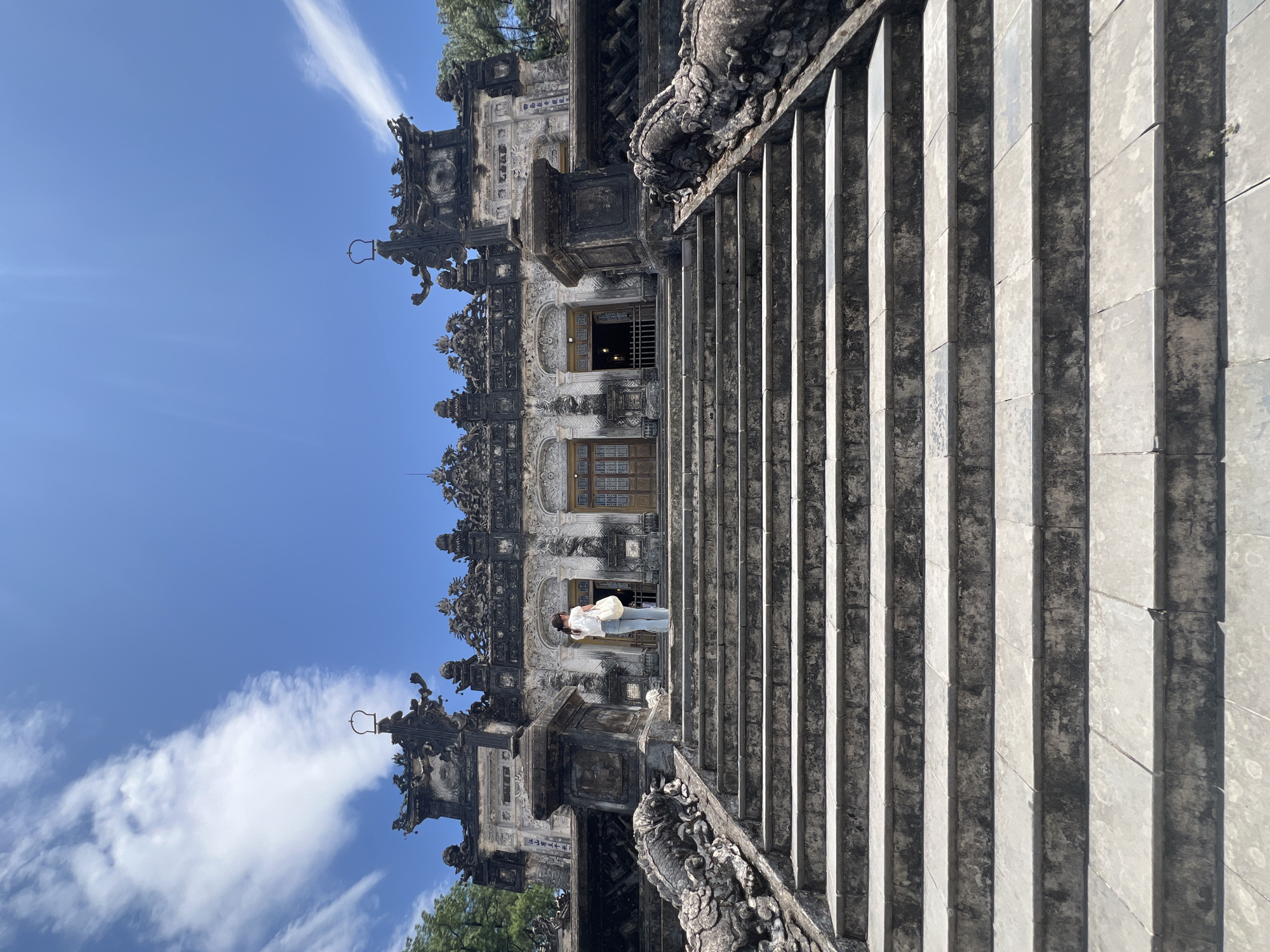 A low-angle, full-shot photograph captures the exterior of a historic building in Thủy Xuân Ward, Vietnam, under a bright, partly cloudy blue sky. A wide, imposing stone staircase, weathered and marked with age, dominates the foreground and leads up to the main structure. On either side of the staircase's lower steps, large, intricately carved stone dragons serve as decorative balustrades, their forms detailed and slightly eroded.
At the top of the stairs, a woman stands facing the building, her back to the camera. She is wearing a white top and light blue jeans and carries a white tote bag over her shoulder. She appears to be looking at the ornate facade of the building. The building itself is grand and elaborately decorated with dark, detailed architectural elements, including multiple tiers of roofs, sculpted figures, and intricate carvings. The entrance to the building features tall, double wooden doors, and archways are visible on either side. The overall impression is one of historical significance and architectural grandeur.