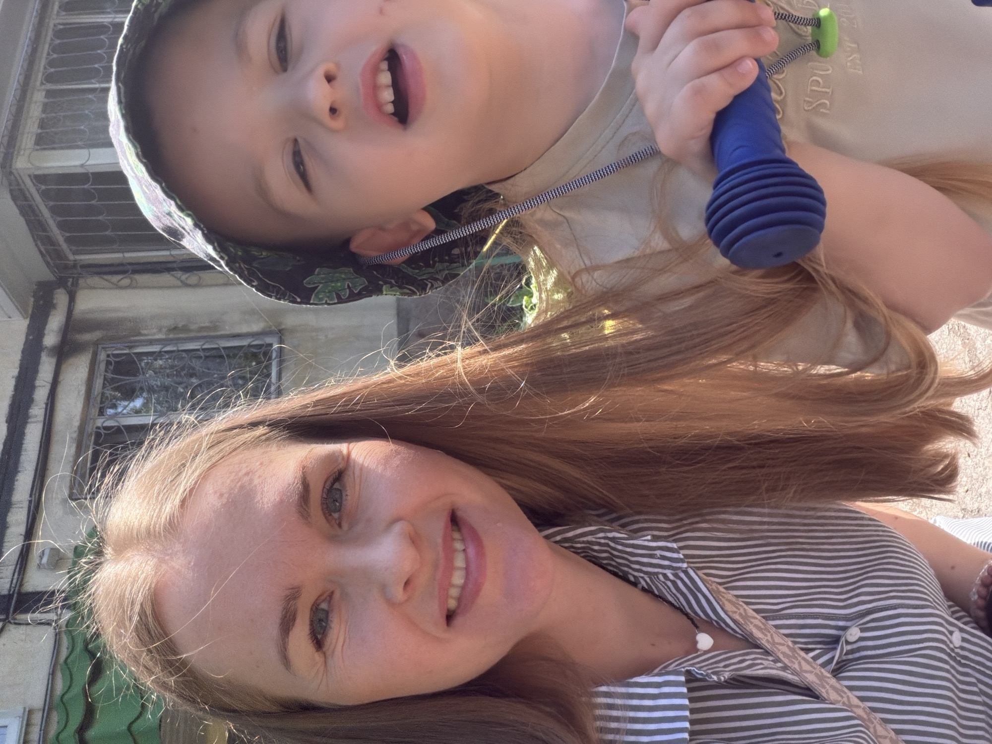 A woman and a young child are taking a selfie outdoors in what appears to be a courtyard or residential area. The woman, on the left, has light brown hair and is wearing a green and white striped button-up shirt. She has a necklace with a white heart pendant. The child, on the right, is wearing a patterned bucket hat and a light-colored t-shirt. The child is holding a blue object with a ribbed texture, possibly a toy or a part of a playground equipment. The background shows a building with windows and a green tiled roof, consistent with residential architecture found in cities like Khabarovsk, Russia. The lighting suggests it is daytime, likely sunny. The overall mood is casual and familial.