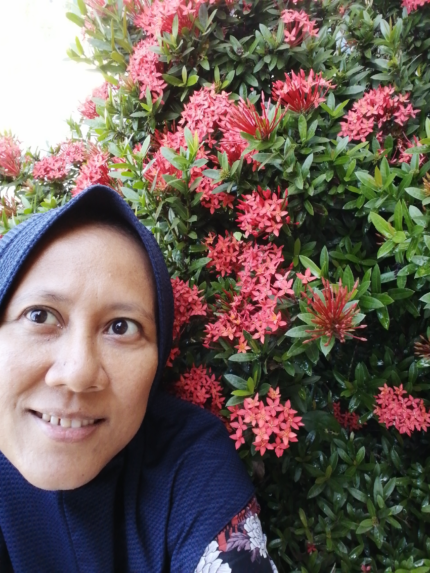 A woman wearing a dark blue hijab and a dark blue top with a floral design on the sleeve is taking a selfie. She is smiling at the camera, and the image is framed with a background of green leaves and small red flowers. The lighting is natural and appears to be outdoors during the daytime. The woman's face is in focus, with the background slightly blurred. The image is a close-up shot of the woman's upper body.