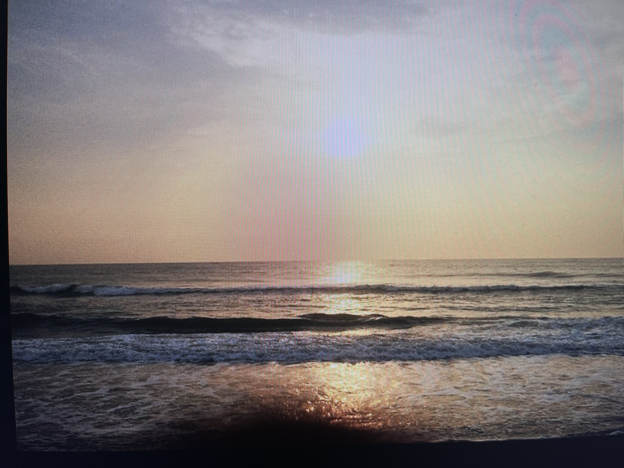 This image depicts a serene beach scene at sunset, likely on the coast of Varna, Bulgaria. The sky is a gradient of soft blues and peaches, with subtle clouds adding texture. The sun, low on the horizon, casts a warm, golden reflection on the calm sea. Gentle waves roll towards the shore, creating lines of white foam against the darkening water. The wet sand in the foreground gleams with the reflected light of the setting sun, hinting at the receding tide. There are no people or distinct structures visible, creating a peaceful and naturalistic atmosphere. The image appears to be captured from a slightly elevated perspective, looking out towards the open sea.