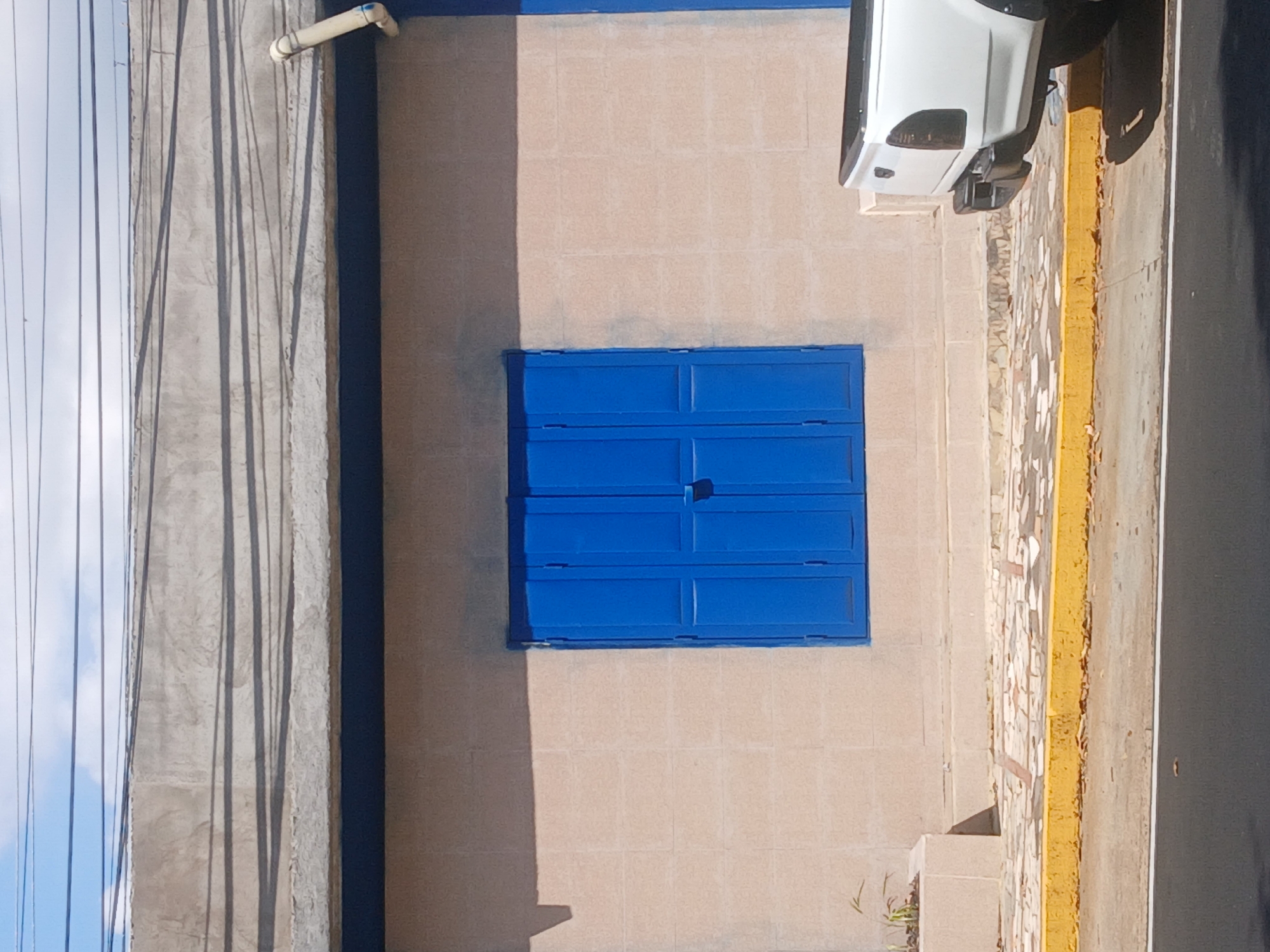 This image depicts a sunlit street scene in what appears to be an urban or semi-urban setting, consistent with Guayana City, Venezuela. The central focus is a building with a light beige, block-patterned facade. Prominently featured on this facade are a set of bright blue window shutters, composed of multiple horizontal and vertical panels, situated roughly in the center of the wall. A narrow strip of blue paint outlines the top edge of the building.
To the upper left of the shutters, a small, light-colored pipe extends horizontally from the wall. On the far left of the frame, a tall concrete structure, possibly a utility pole or an adjacent building's wall, is visible, marked with several long, dark shadows cast by overhead wires. Beyond this structure, a clear, bright blue sky is interlaced with thin utility lines.
The right side of the image shows a street area. A paved surface, with a mosaic-like texture in places, leads to a bright yellow curb. Beyond the curb is a dark asphalt road. The rear section of a white pickup truck, including its taillights and a black bumper, is parked alongside the yellow curb in the upper right portion of the frame. Small patches of green vegetation are visible near the base of the building and along the curb.
The sharp, distinct shadows cast by the building and the overhead wires strongly indicate a sunny day, likely during midday or early afternoon. No people or visible text are present in the image.