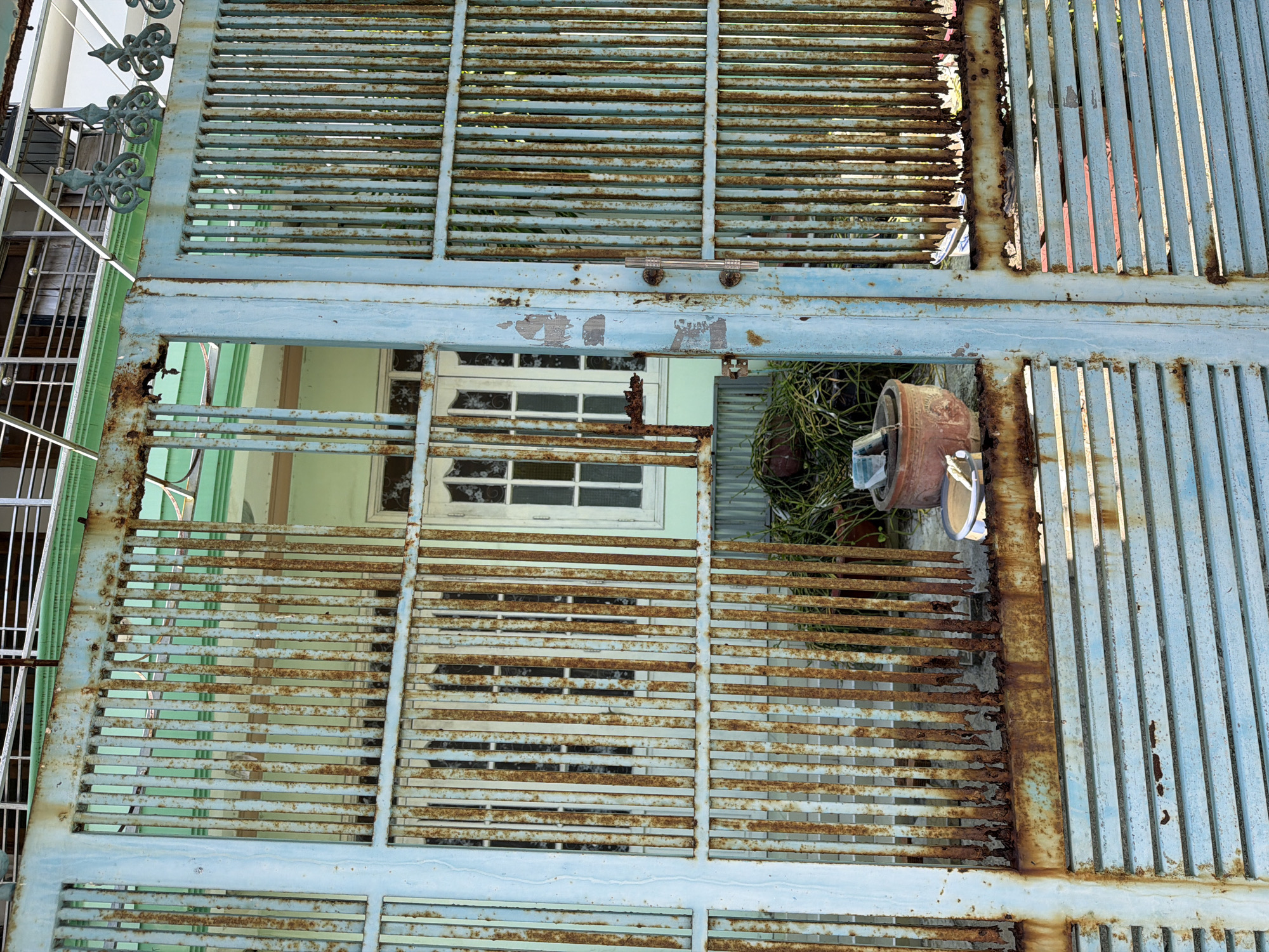 The image depicts a weathered, pale blue metal gate with significant rust and peeling paint, located in a residential area of Nha Trang Ward, Vietnam. The gate is partially open, revealing a glimpse of a light green building with white-trimmed windows behind it. In the foreground, at the base of the gate, there is a terra cotta pot containing green foliage and a small, round, light-colored bucket with some white material inside. The gate's vertical bars are heavily corroded, and some at the bottom appear broken or significantly weakened. The overall scene conveys a sense of age and disrepair.
