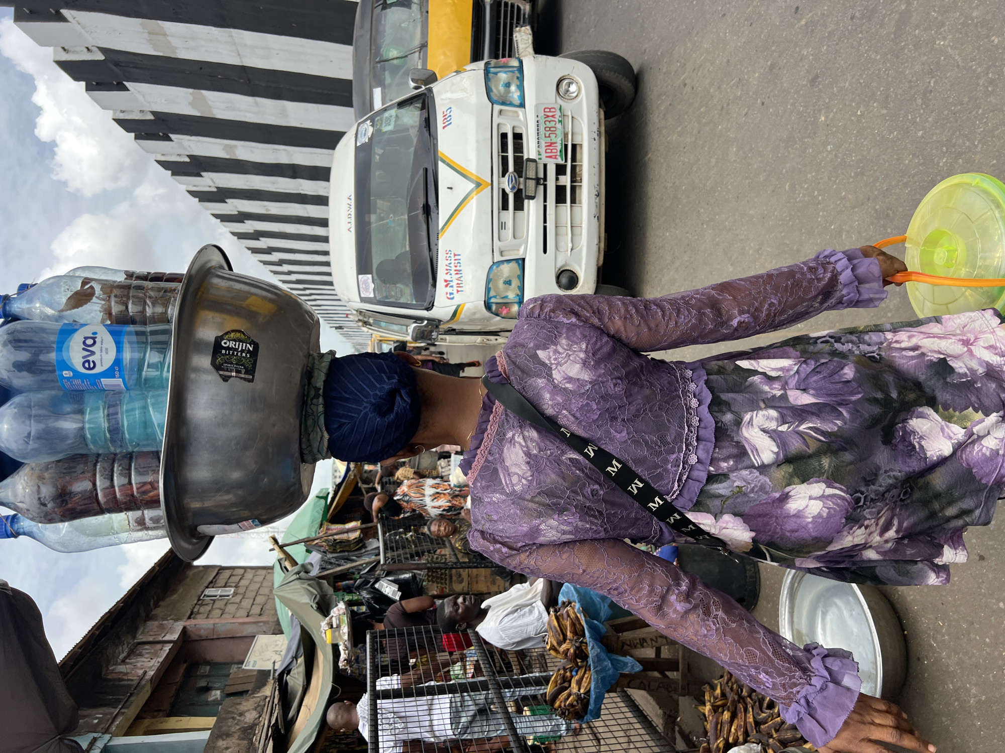 The image shows a woman carrying water bottles in a metal bowl on her head, standing on a street in what appears to be Nkpor, Nigeria, based on the vehicle license plate. She wears a purple dress and has a strap across her shoulder. In the background, there's a white van with "G.M. MASS TRANSIT" written on it and license plate "ABN-583XB." There's also a black and yellow taxi visible, along with a bustling street market scene. Bananas are seen in a cage. A bowl can be seen in the woman’s hand. A bridge is visible.