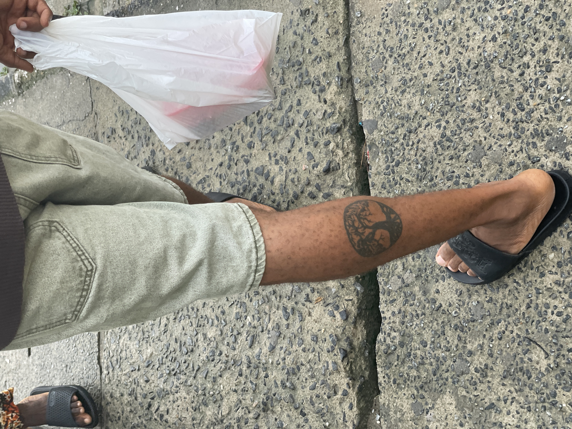 The image depicts the lower extremities of two individuals on a textured paved surface in Ado, Nigeria. The primary subject, positioned centrally, wears light grey or khaki shorts. Their right lower leg, visible on the left side of the frame, exhibits a dark, circular tattoo depicting a tree with exposed roots and branches. This individual wears black slip-on sandals on both feet. Their right hand, at the upper right, holds a crinkled white plastic bag, partially revealing red or pink contents. The second individual is partially visible on the far left, showing their left foot, also wearing a black slip-on sandal. A patterned fabric is visible around their ankle. The ground consists of a rough, grey aggregate pavement with embedded dark pebbles, featuring a distinct horizontal crack or gap running across the mid-section of the frame. A small patch of green vegetation is discernible in the upper right background.
