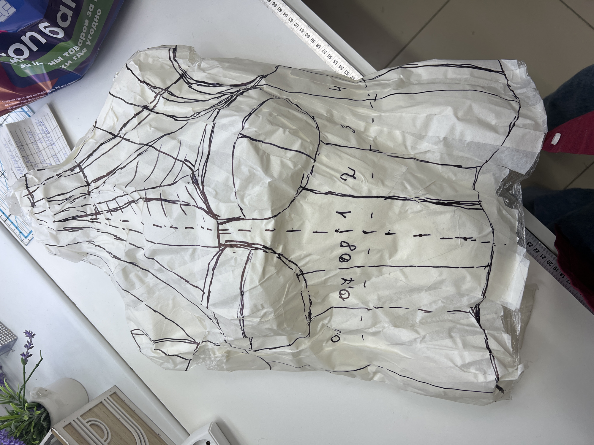 Here's a detailed description of the image:
**General Description:** The image shows a crumpled piece of paper with drawn lines and markings, resembling a pattern for clothing or garment construction. It is placed on a white table surface.
**Subjects and Objects:**
* **Pattern:** The central subject is the white, crumpled paper sheet with dark lines indicating the pattern. It appears to be a bodice pattern, possibly for a dress or top, with distinct curves for the bust area.
* **Ruler:** A metal ruler is partially visible near the top edge of the pattern.
* **Other Items:** There are other items in the periphery, including a bag with Russian text, a small decorative plant, and other miscellaneous objects on the table.
**Environment:**
* **Setting:** The image appears to be taken in a workshop or studio setting. The white table suggests a clean workspace for design or crafting.
**Text and Markings:**
* **Handwritten Notes:** Handwritten text in Russian is visible on the pattern.
**Overall Impression:**
The image conveys a sense of the design and construction process in fashion or garment making. It focuses on the technical aspects of pattern creation and tailoring.