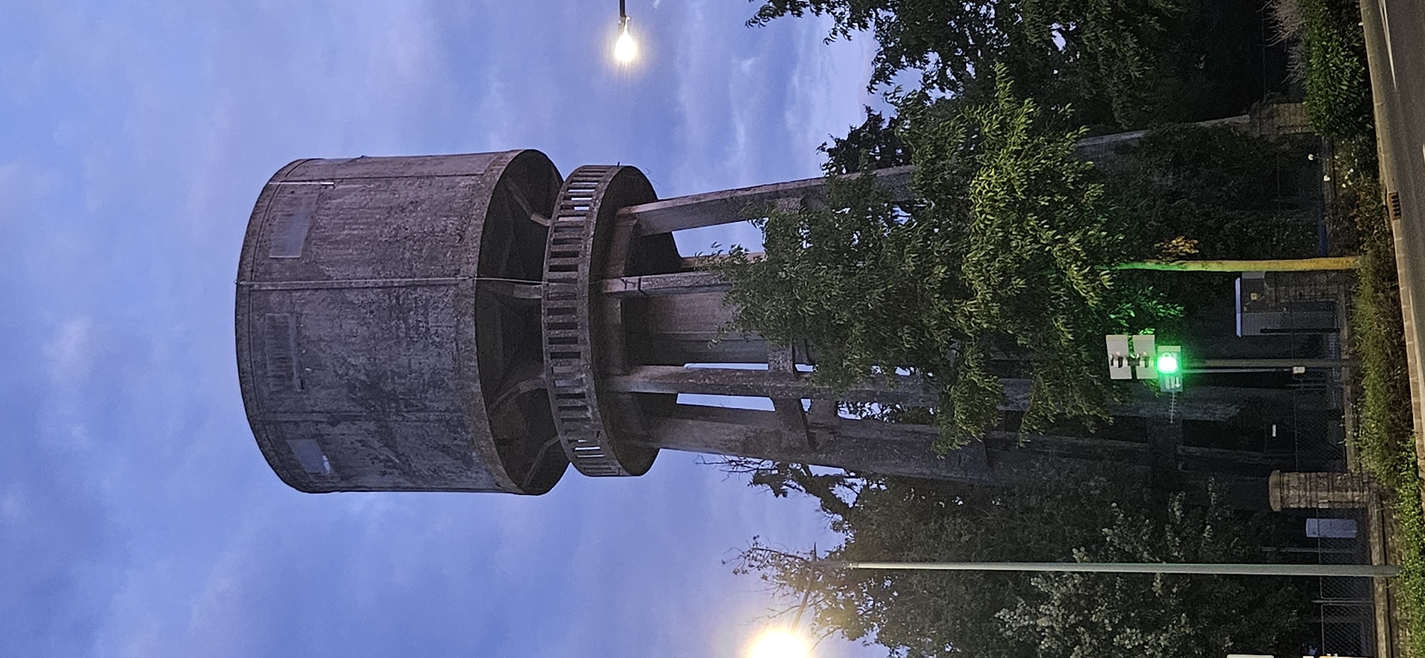 A large, cylindrical, concrete water tower stands tall against a twilight sky. The tower has a weathered, rough texture and is supported by four thick, trụ cột legs. A circular balcony with a balustrade encircles the base of the cylindrical tank. Several rectangular windows are visible near the top of the tank. Surrounding the base of the tower are various trees and foliage, with some greenery obscuring the lower portions of the structure. In the foreground, a street light illuminates the scene, and a traffic signal displaying a green light is visible on a pole to the left of the tower. The overall impression is one of an industrial structure set amidst a natural environment as dusk settles. This water tower is located in Hévíz, Hungary.