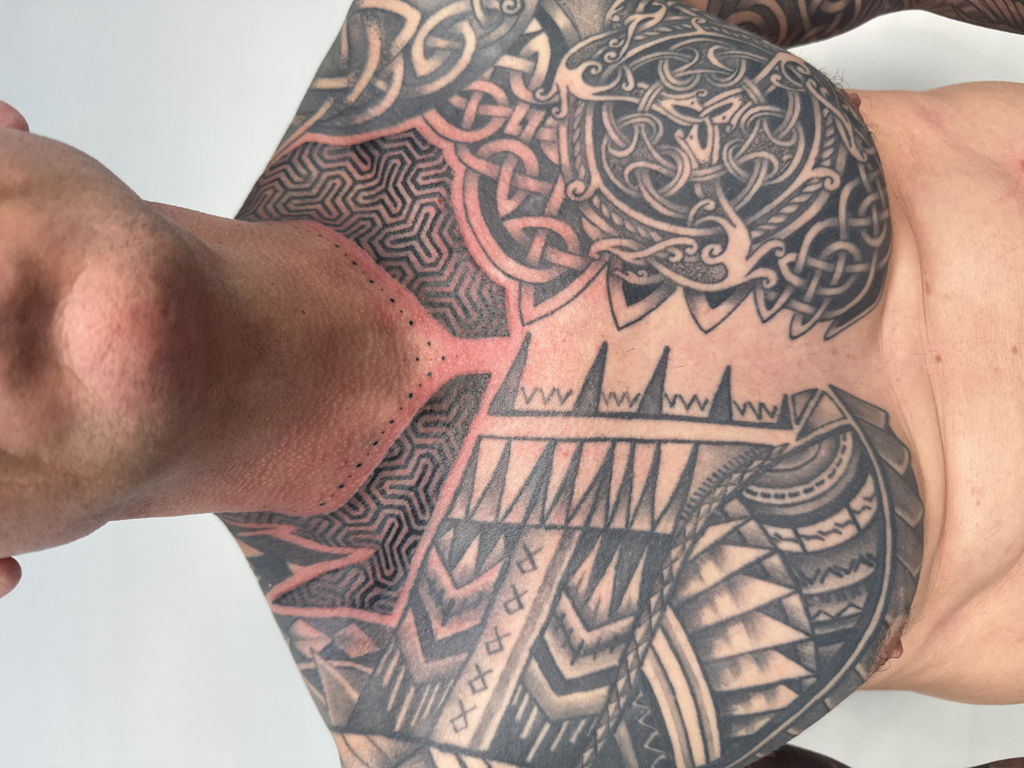 This media file captures a close-up, top-down view of a person's heavily tattooed chest, shoulder, and upper arm, presented against a plain white background. The subject is likely lying down, with their head and neck partially visible on the left, facing upwards.
The extensive tattoo work covers a large area of the upper torso, showcasing diverse patterns. Intricate Celtic knotwork designs in dark grey and black shading are prominent on the upper chest and shoulder. Adjacent to these, closer to the neck and collarbone, are geometric maze-like patterns with bold black lines. These distinct design areas are often bordered by sections of reddish skin, suggesting the tattoos are either fresh, recently worked on, or in the process of healing. Tribal-inspired motifs, characterized by sharp angles, chevrons, and thick lines, extend across the lower chest and wrap around the side, some featuring lighter grey shading.
The skin around the neck and upper chest appears red and slightly irritated, indicative of recent tattooing. Small black dots, possibly from a stencil or a dot work technique, are visible along the neck line. The person's facial features are largely out of frame, so no emotions can be discerned. The lighting is bright and even, effectively showcasing the detail and texture of the body art. The plain background suggests an indoor, professional setting, such as a tattoo studio in Sofia, Bulgaria. There is no visible text in the image.