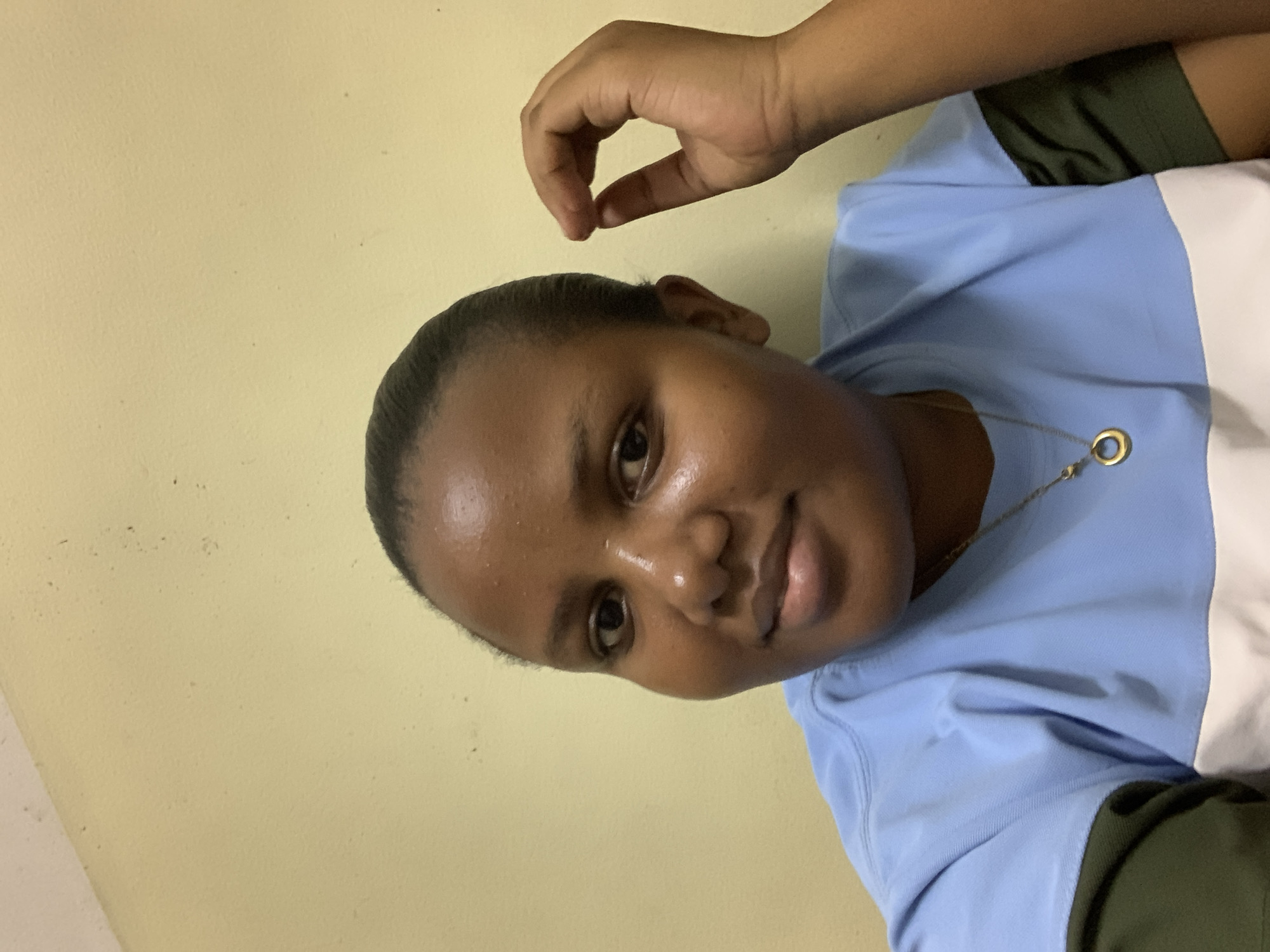 A person is posing for a close-up shot. The individual has dark skin, short dark hair, and is wearing a light blue and white shirt with olive green sleeves. A gold necklace with a circular pendant is visible. One hand is positioned above the head. The background is a plain, light yellow wall. There is no information to support that this photo was taken in Dar es-Salaam, Tanzania.