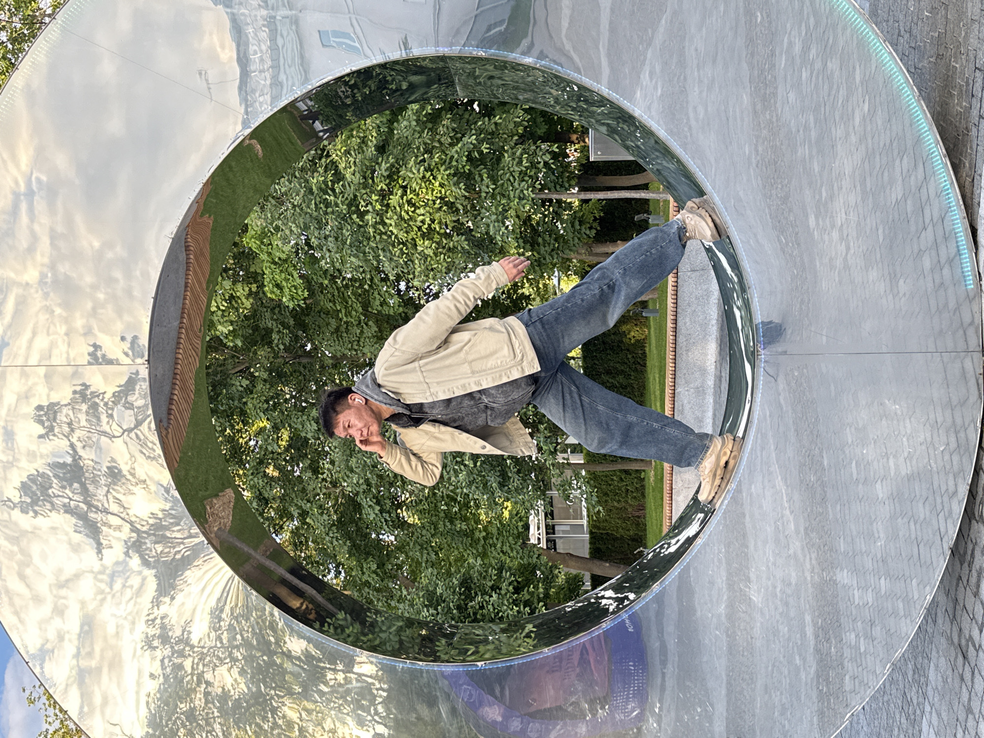 A man is captured in a dynamic pose, balancing on the inner edge of a large, highly reflective circular structure outdoors in Moscow, Russia. The man, with dark hair and wearing a beige jacket over a darker top, blue jeans, and light brown shoes, has one hand near his ear, possibly adjusting an earbud. He appears to be leaning back, using the curved surface as a prop for his pose.
The dominant feature of the scene is the massive, chrome-like circular art installation or tunnel. Its polished surface creates a distorted, mirror-like reflection of the surroundings, showing lush green trees, bright white clouds, and distant buildings. The reflection also hints at a clear, sunny daytime setting. Along the inside perimeter of the circular structure, a strip of glowing, teal-colored light is visible, adding a futuristic touch. The outer part of the circular structure appears to be made of a textured grey material, resembling paving stones or concrete.
Through the center of the ring, an abundance of green foliage and trees suggests a park or urban green space. The overall impression is one of playful interaction with public art, captured in bright daylight. Some unclear, distorted white text is faintly visible reflected on the lower, purplish-blue section of the reflective structure.