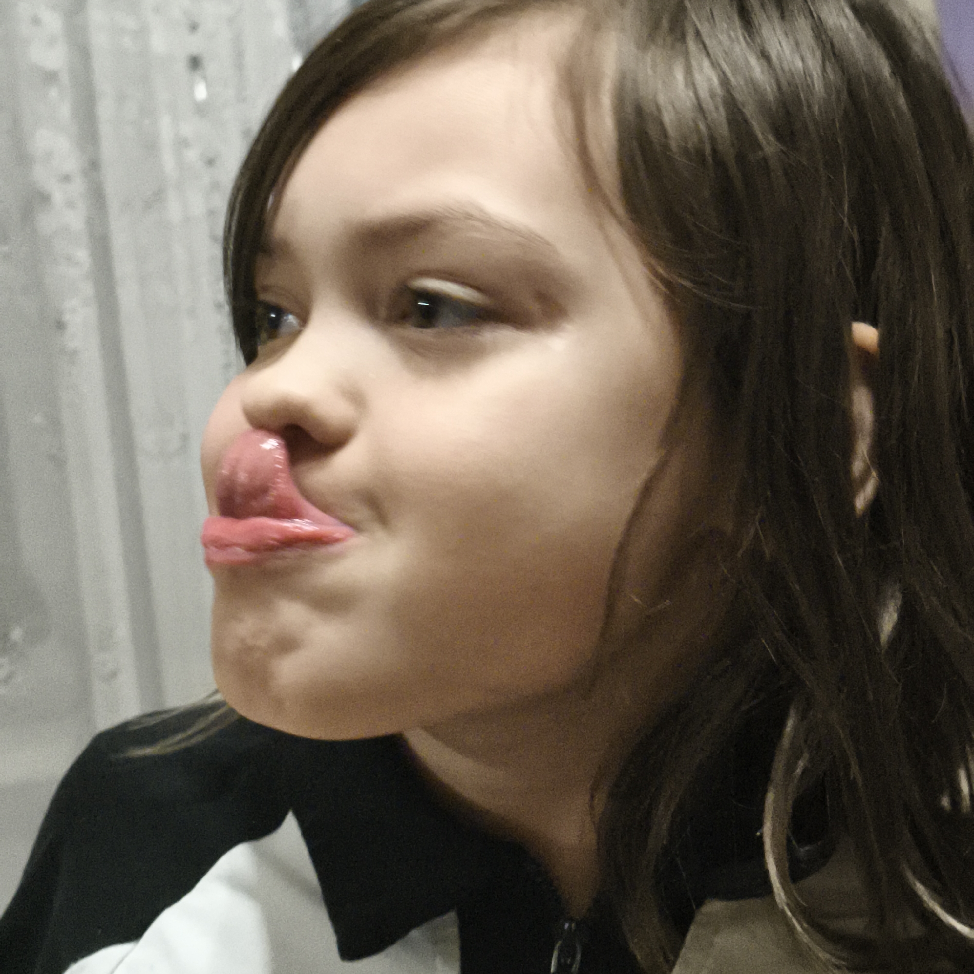 A young girl with dark hair is shown in a close-up shot. She is sticking her tongue out, with her lips pursed and nose slightly scrunched. She is wearing a black and white shirt. The background is slightly blurred, showing a patterned curtain. The location is Unknown, Unknown.
