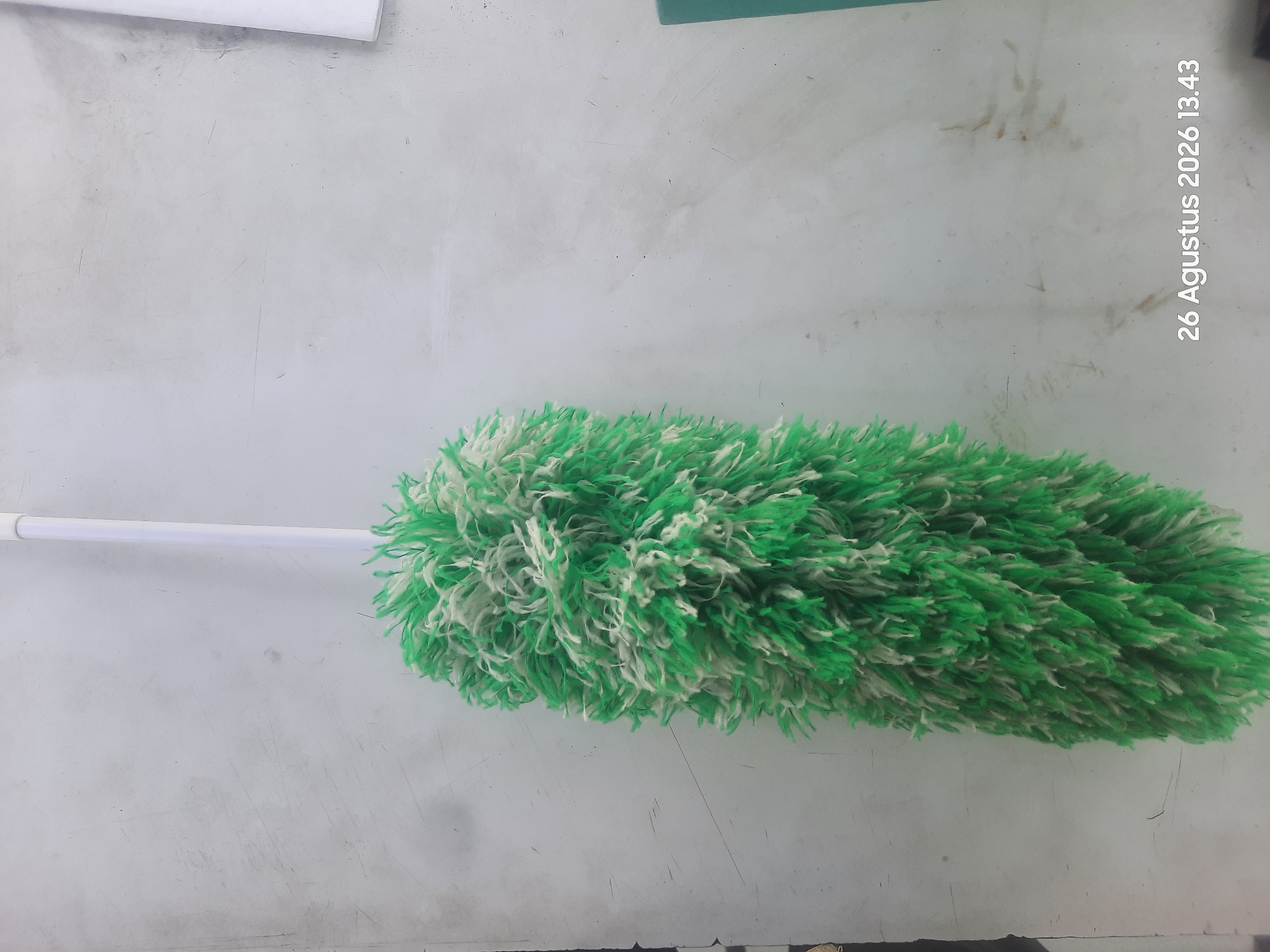 A green and white duster is hanging vertically against a textured, light-colored wall. The duster's fluffy head is at the bottom of the frame, and its white handle extends upwards and out of view. The wall has visible scuff marks and some faint brown smudges. In the bottom right corner, a timestamp reads "26 Agustus 2026 13.43". The location is Gending, Indonesia.