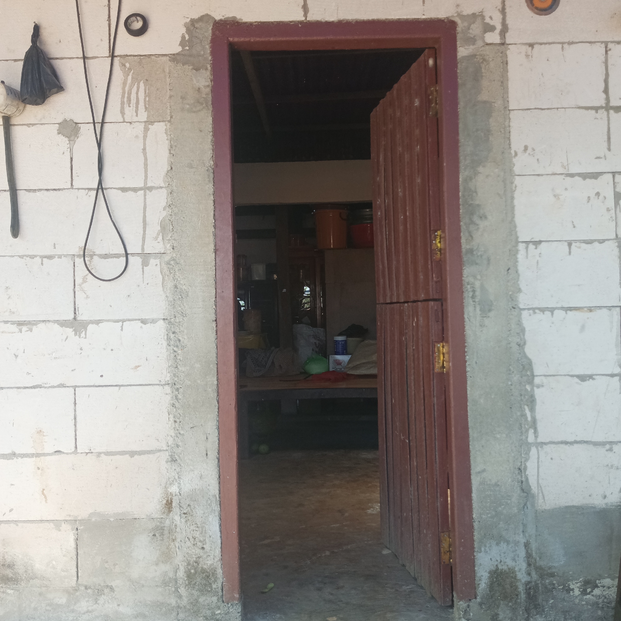 The image shows an exterior view of a building under construction, possibly in Tumampua, Indonesia. The walls are made of white cinder blocks with visible mortar lines. A doorway with a dark red metal frame is partially open, revealing the interior. The door itself is also dark red and appears to be made of corrugated metal. On the exterior wall to the left of the door, a black plastic bag is hanging, along with two dark electrical wires. One wire is looped and hanging loose, while the other is taut and runs vertically. A black circular object is mounted on the wall near the top left corner. The ground in front of the doorway is a concrete surface, and the interior of the building appears cluttered with various objects, including containers, bags, and what looks like furniture. The lighting suggests it is daytime.