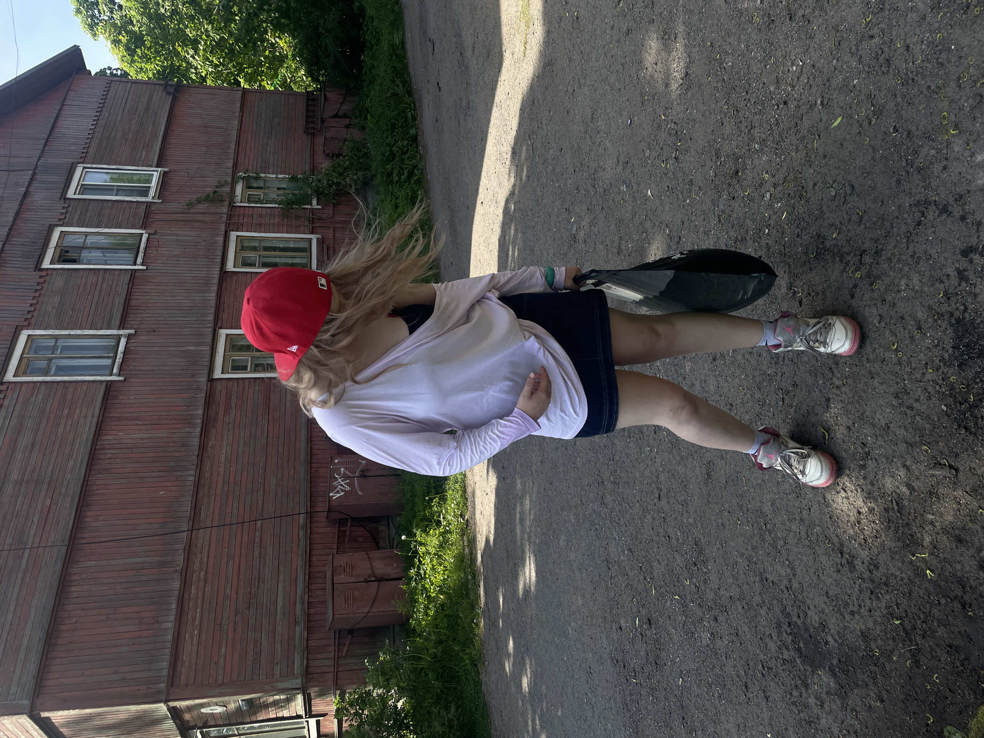 The image depicts an outdoor scene in Priozersk, Russia, under daylight conditions. A single individual stands on a dirt and gravel path in front of a two-story wooden building.
The individual, positioned centrally, has long blonde hair partially covered by a red baseball cap featuring a white logo on its left side, consistent with the "New Era" brand. They are wearing a light pink long-sleeved shirt, which is draped to reveal a black garment underneath the upper torso. A dark denim mini-skirt, white socks, and white sneakers with pink accents complete their attire. The person's head is tilted downwards and to their left, with their hair appearing in motion, blowing towards their right. A black bag is held in their right hand, resting by their side, while their left hand is placed on their abdominal/hip area.
Behind the individual, a weathered two-story building with a reddish-brown wooden facade is visible. The building features vertical plank siding on its upper and lower sections, with a band of horizontal planking across the middle of the second story. Multiple rectangular windows with white frames are present across both floors. On the ground level, to the left of the individual, two dark reddish-brown metal utility boxes or lockers are located at the base of the building, connected by a chain. Graffiti, appearing to be "DAS," is etched onto the rightmost utility box. A thin, dark cable or wire extends diagonally downwards across the upper left portion of the building's facade.
The ground consists of an uneven dirt and gravel path, interspersed with small green plants, scattered leaves, and debris. Shadows are cast on the path, indicating direct sunlight. Lush green foliage and trees are visible on the right side of the frame and around the base of the building, particularly on the left.
