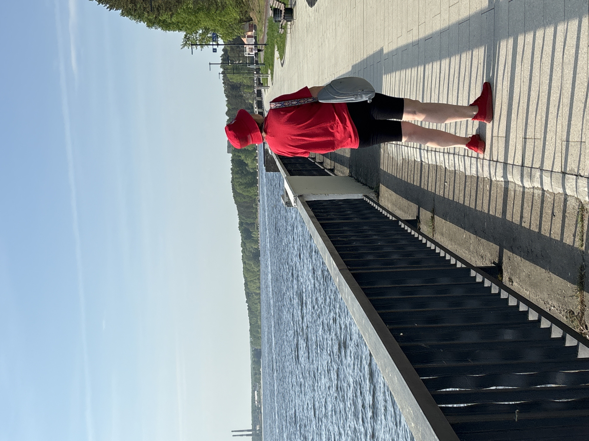 A person dressed in a red t-shirt, red hat, and red sneakers with black shorts stands on a paved path alongside a waterfront staircase. The person also has a gray bag on their shoulder. The staircase descends towards a body of water. Across the water, there is a line of green trees. A bright blue sky is visible in the background. The location is Chaikovsky, Russia.
