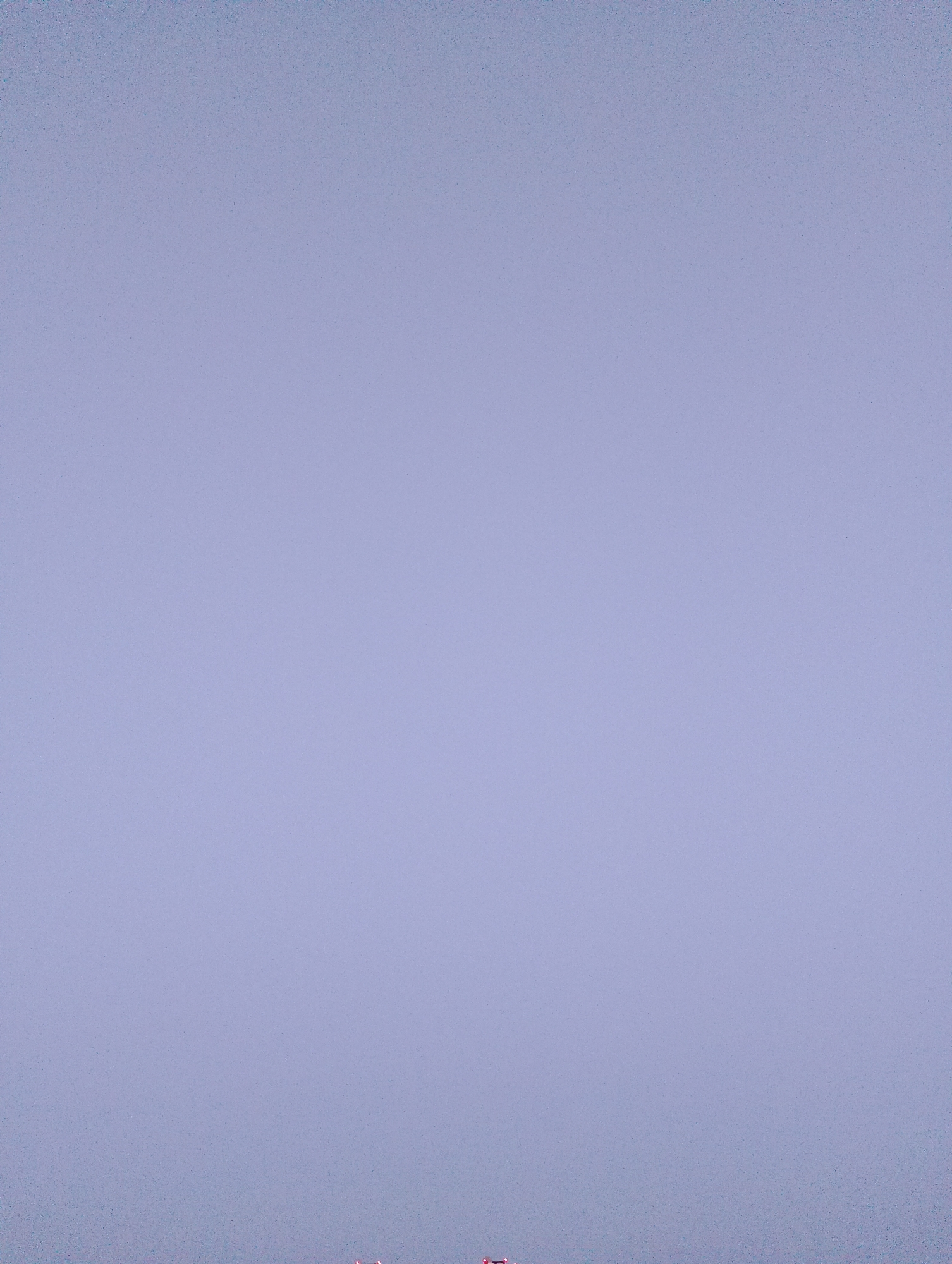 The image displays a hazy, pale blue sky with some subtle texture from atmospheric dust or light. At the very bottom edge of the frame, a faint line of red lights is visible. These lights are indistinct and appear to be part of a distant structure or cityscape, possibly representing the illuminated windows or rooftop lights of buildings. The overall impression is one of a broad, overcast sky with a hint of urban presence at its lower boundary. The specific location in Viciebsk, Belarus, cannot be determined from the visual information present in the image.