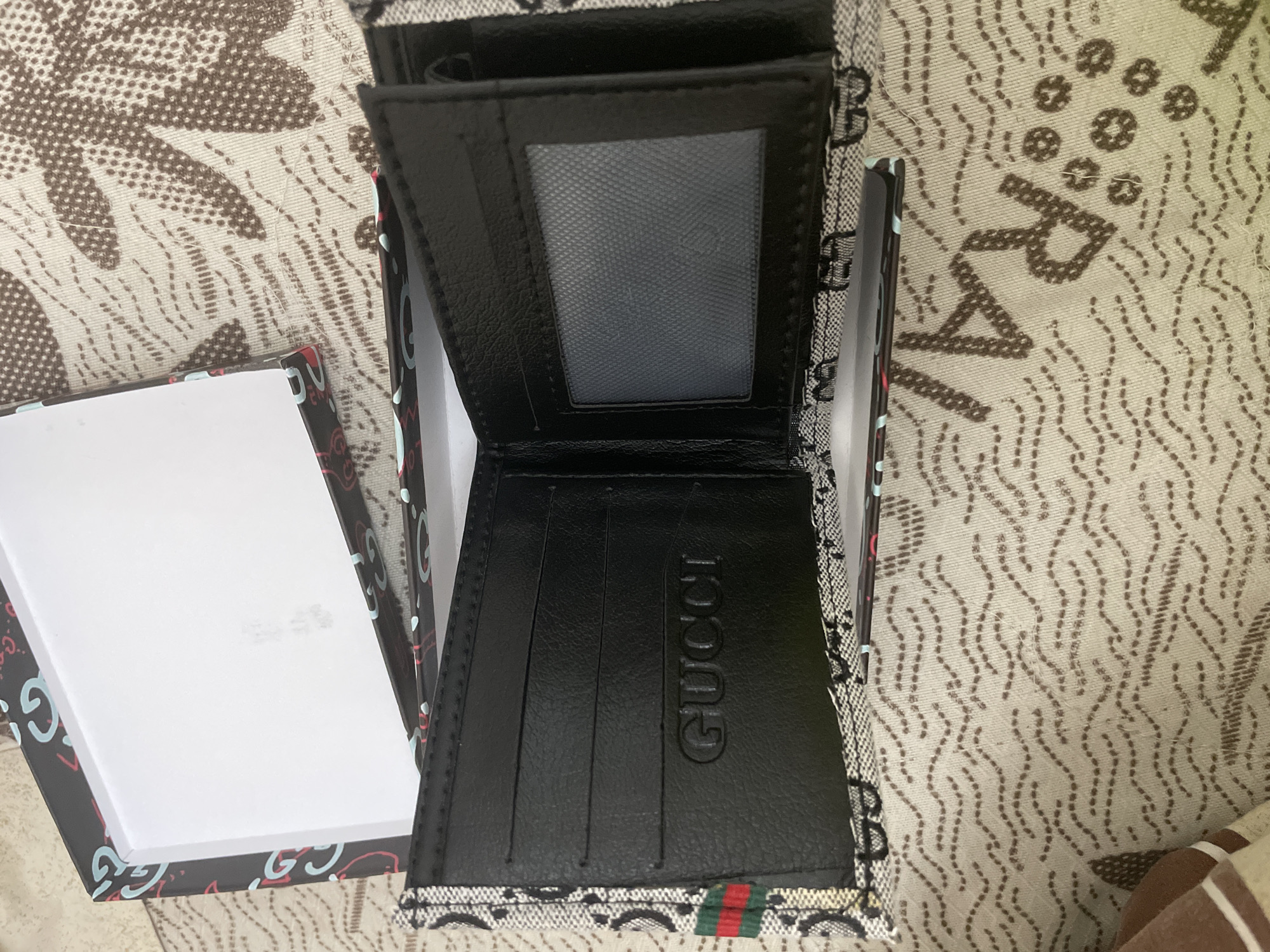 An open black bifold wallet is centrally displayed, resting within its gift box on a patterned fabric surface. The wallet's interior is made of a smooth, black leather-like material, featuring several card slots and a transparent ID window with a textured gray surface. The word "GUCCI" is clearly embossed on one of the inner flaps of the wallet. A distinctive red and green striped band, a common design element for the brand, is visible along the bottom right exterior edge of the wallet.
The gift box is black with a repeating pattern of light blue and red "GUCCI" logos. The inside of the box is white, with some faint, illegible text or markings on its surface. The exterior edges of the wallet also feature a subtle "GG" monogram pattern.
The background consists of a light-colored fabric, likely a tablecloth or bedding, adorned with abstract brown patterns. These patterns include wavy dotted lines and clusters of small circles, with some shapes resembling stylized letters such as an 'R'. The scene is indoors, under artificial or diffuse natural light. No individuals are present, and the image primarily focuses on the presentation of the wallet in its packaging. While the context suggests the location is Ihtiman, Bulgaria, there are no specific visual cues within the image to confirm this geographical detail.