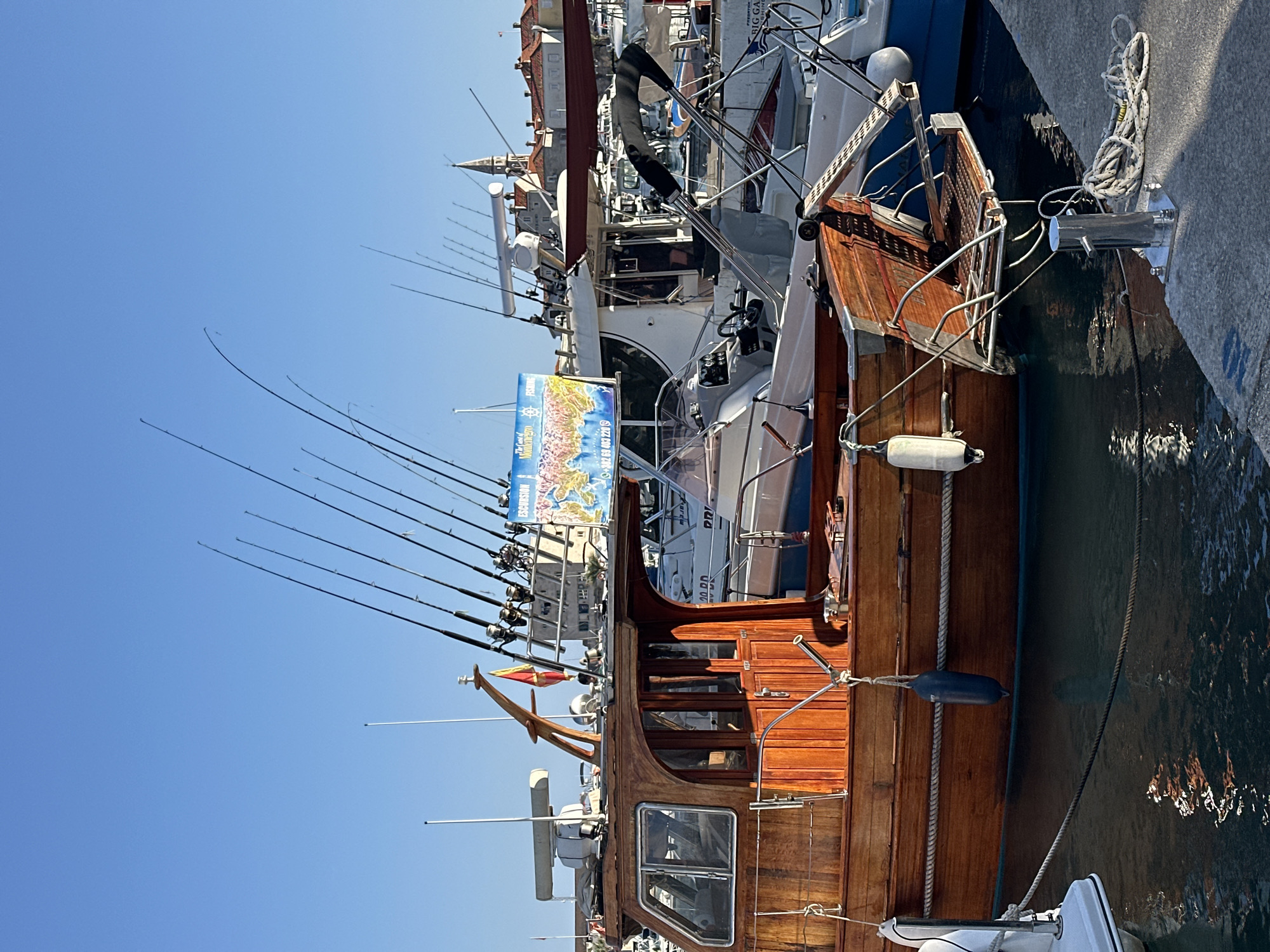 This image, rotated clockwise by 90 degrees, captures a bright, sunny day at a marina, likely in Budva, Montenegro, given the visual cues and provided context.
The foreground is dominated by a beautifully varnished wooden boat, appearing to be a fishing or pleasure craft, docked in clear blue water. Numerous fishing rods are neatly mounted along its side, indicating its purpose. The boat features a wooden cabin with windows and is secured by ropes, with fenders visible along its hull.
In the mid-ground, other boats are docked, including a white fiberglass yacht with visible rigging and radar equipment. Behind the boats, the architecture of a coastal town is visible, featuring buildings with red-tiled roofs and a prominent church steeple rising into the clear blue sky.
A colorful banner or sign is displayed on one of the white boats. Visible text on this sign includes "EXCURSION" and "The Coast of MONTENEGRO," along with a phone number "062 83 003 221" and a logo depicting a fish and a ship's wheel. The bright sunshine and cloudless sky indicate fair weather.